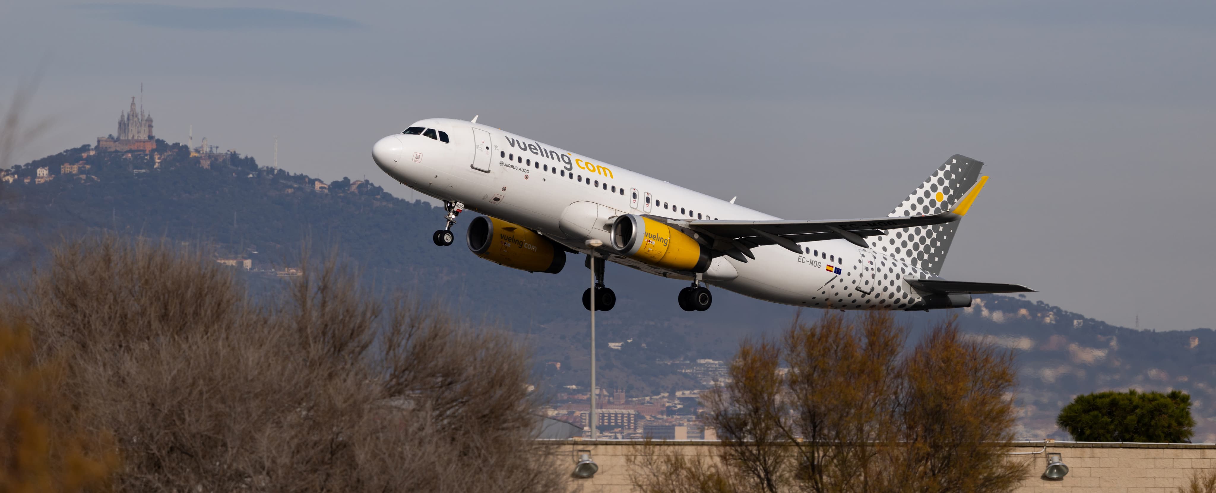 A Vueling Airlines plane takes off against a mountainous backdrop with a blurred landscape in the foreground.