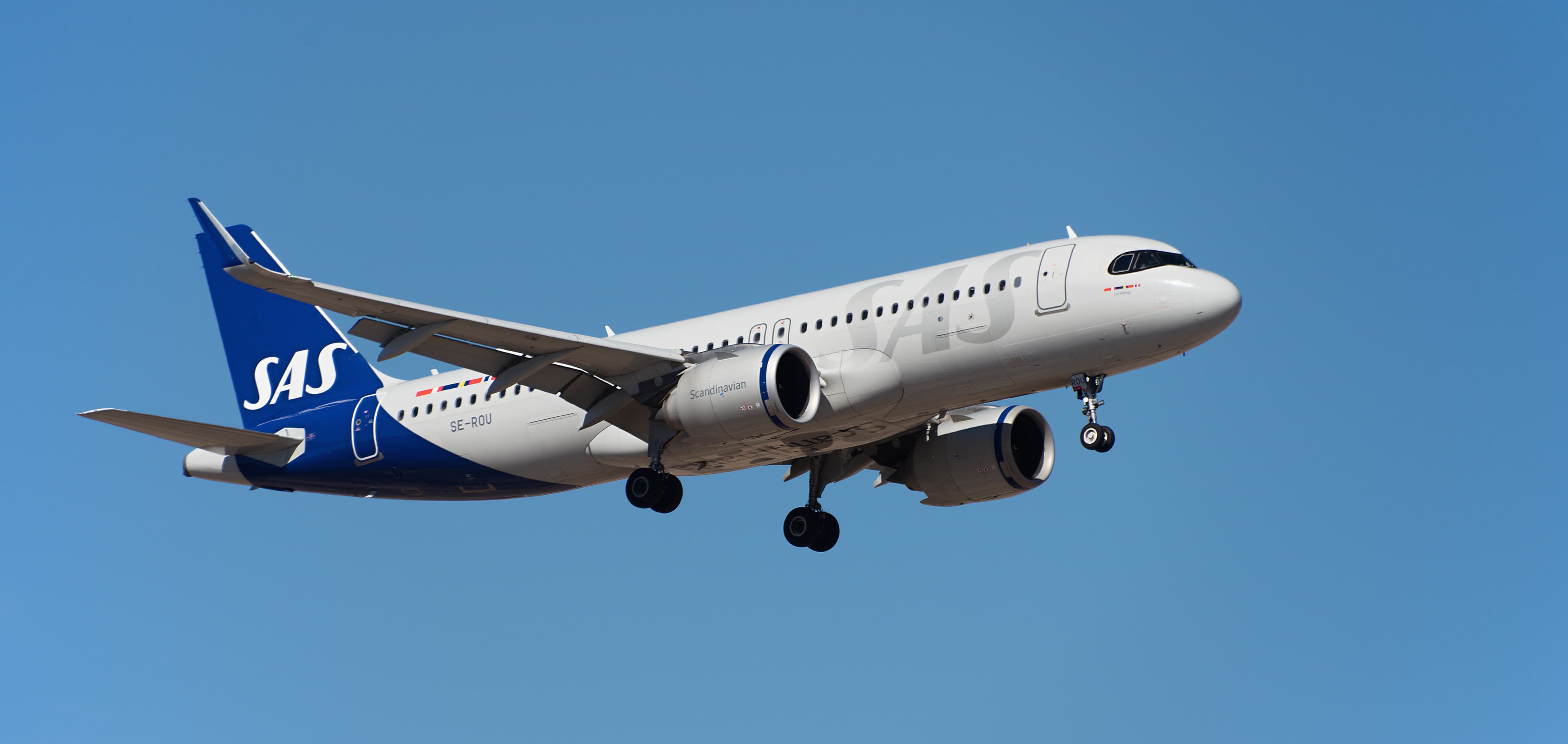 Airplane with blue tail and visible landing gear flying against a clear blue sky.
