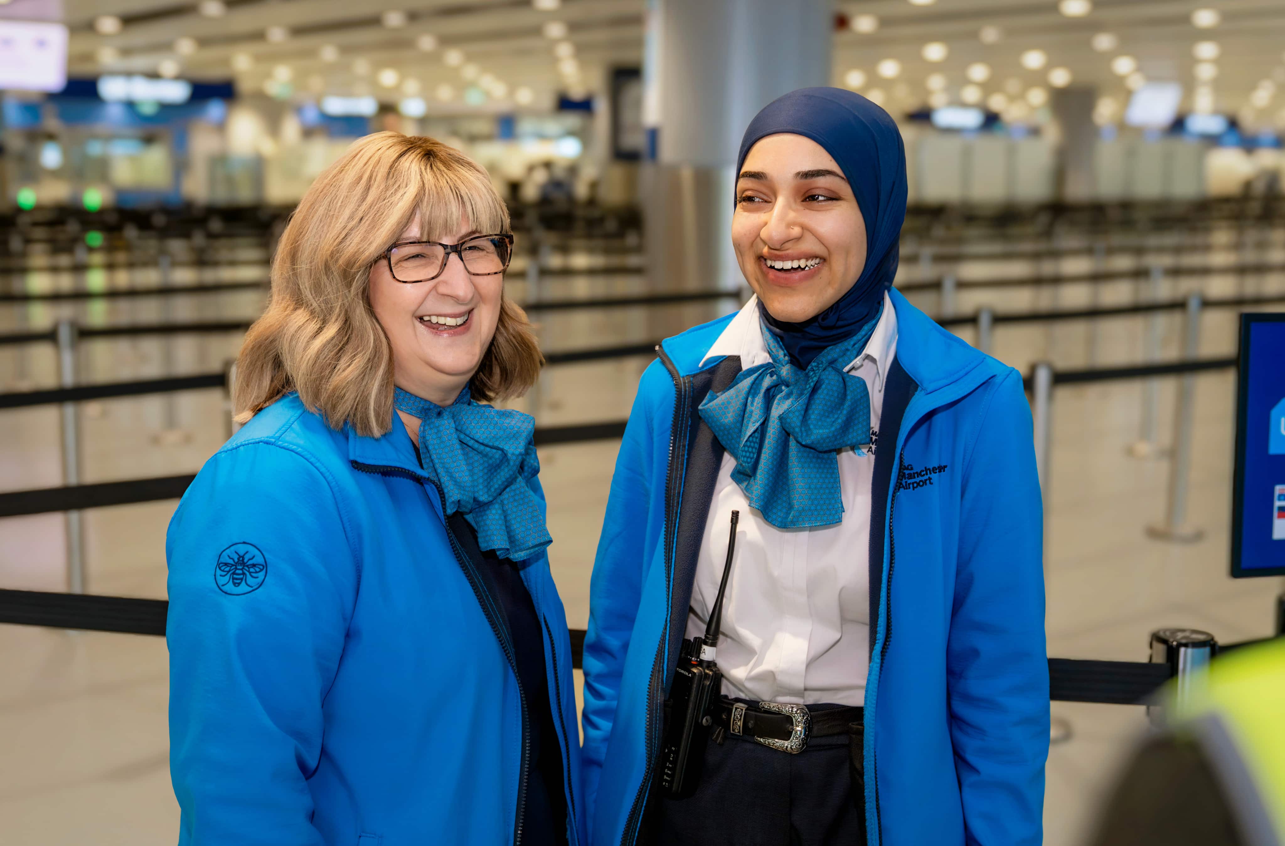 Two airport staff in blue uniforms, smiling and chatting at a terminal with blurred background.