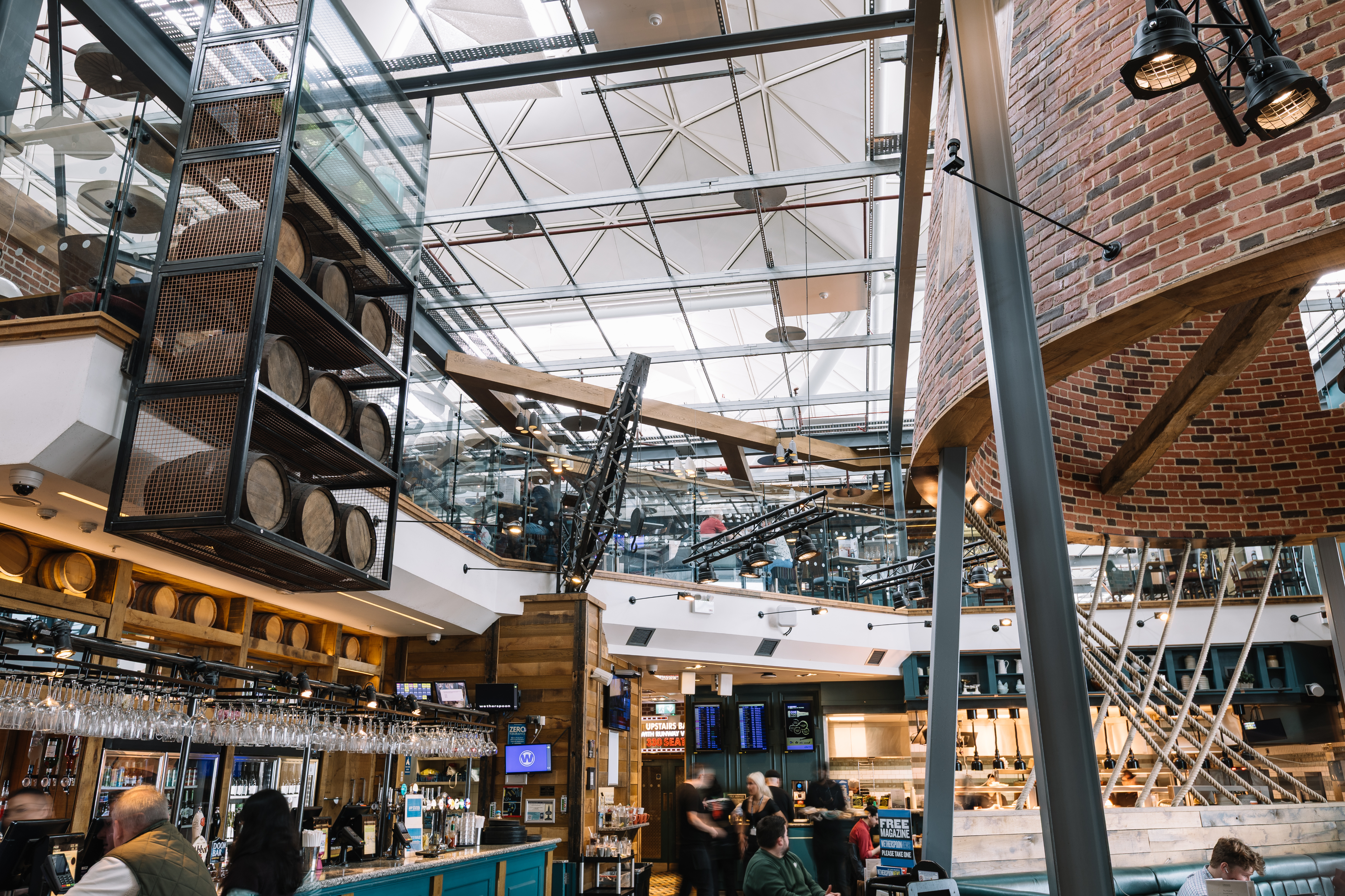 Modern, spacious bar interior with wooden barrels, glass panels, exposed beams, and a lively atmosphere. People are sitting and walking around.
