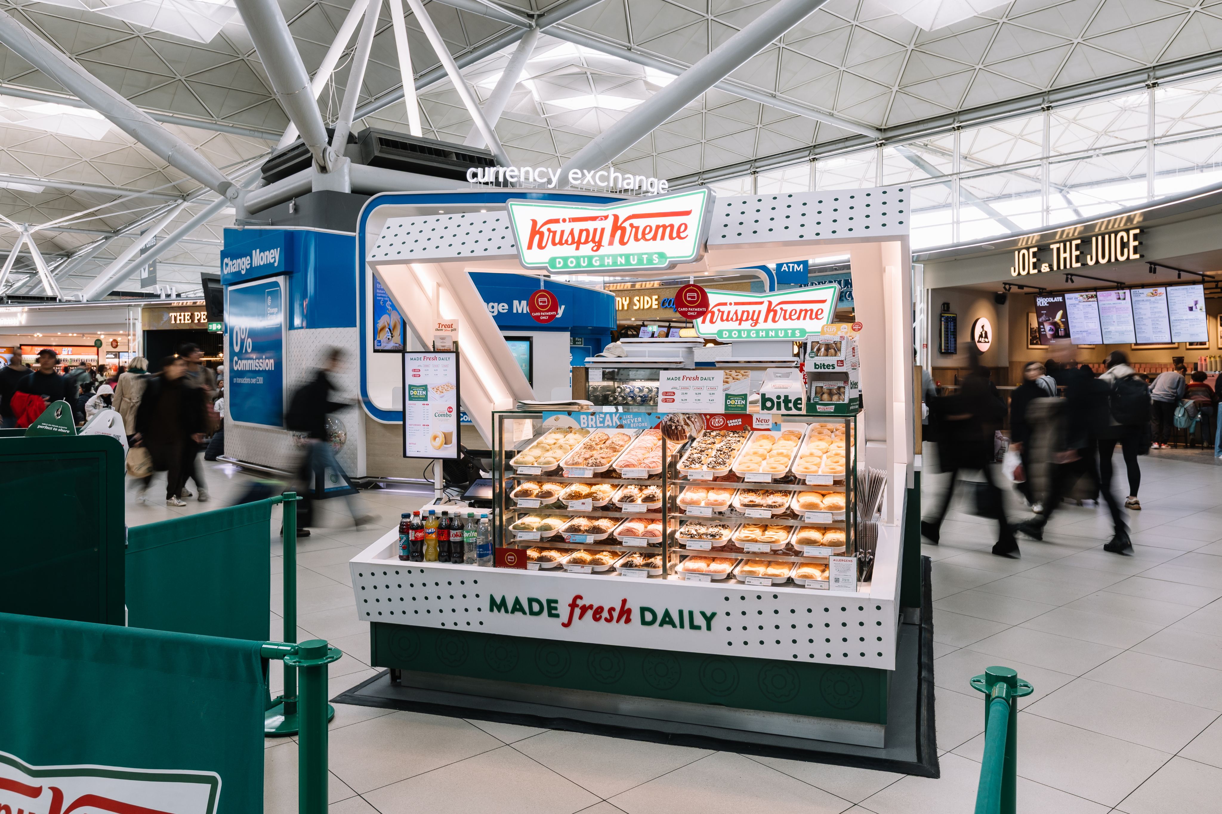 Krispy Kreme kiosk in a busy airport, displaying assorted doughnuts and drinks. Currency exchange and Joe & the Juice in the background.