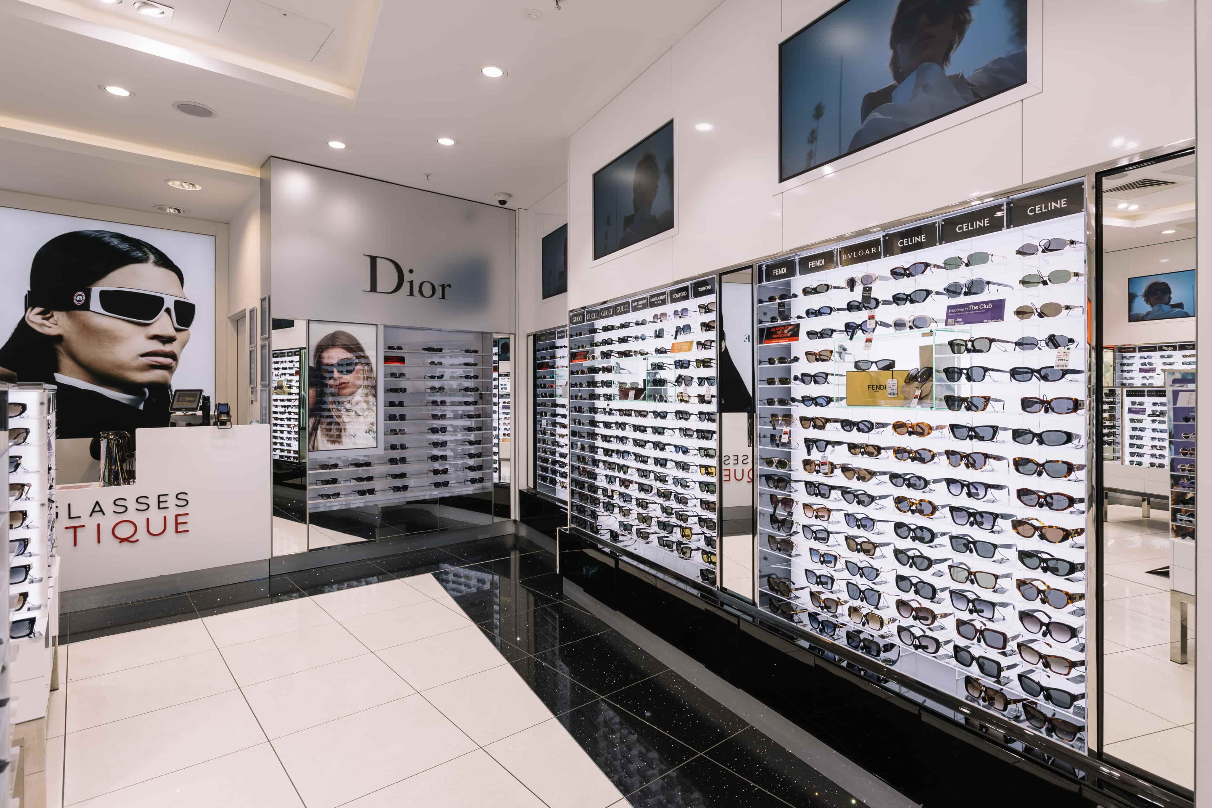 Sunglasses store interior with glass shelves displaying various designer brands like Dior, Gucci, and Celine, and large model posters.