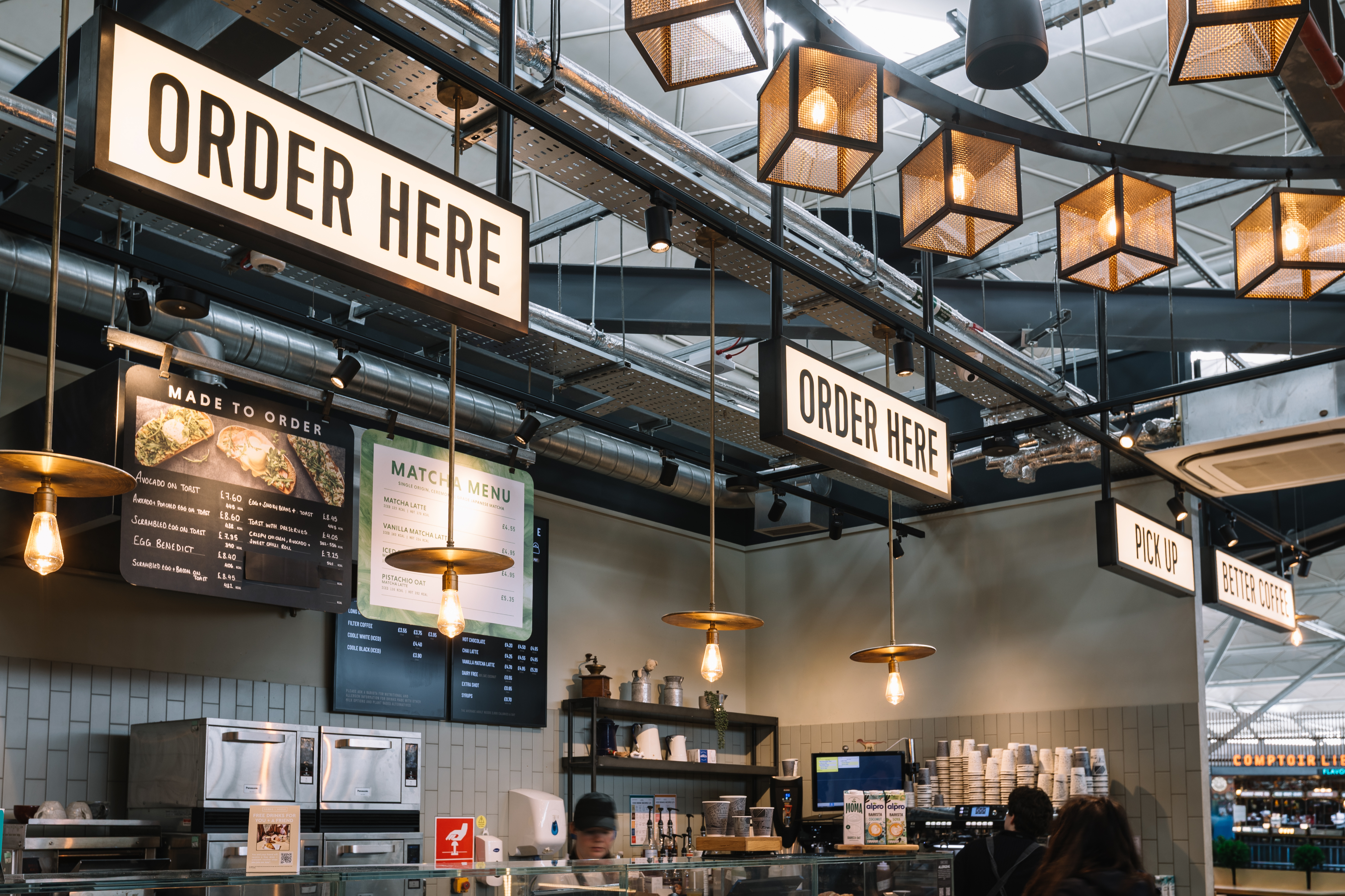 Cafe interior with signs reading "Order Here" and "Pick Up," hanging lights, menu boards, and a counter with kitchen equipment and cups.