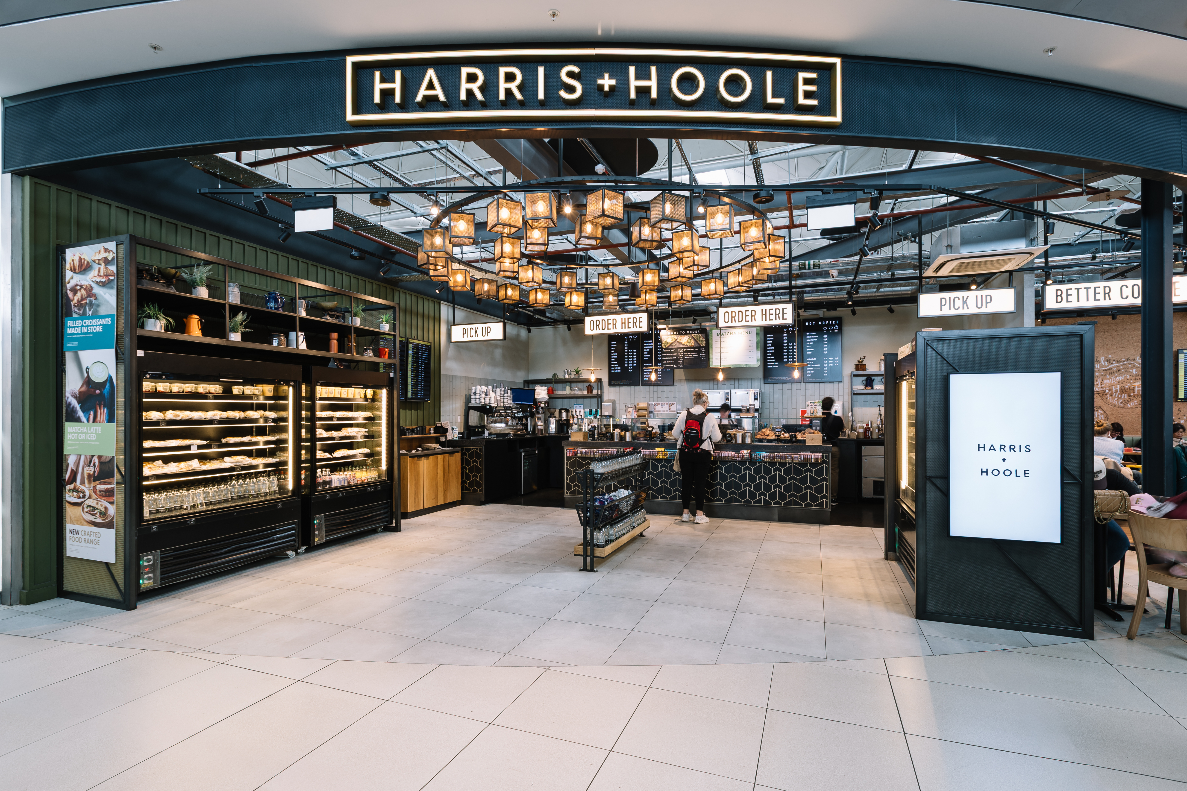 Modern coffee shop with pastry displays, hanging lights, and order counters. Signs read "Harris + Hoole," "Order Here," and "Pick Up."
