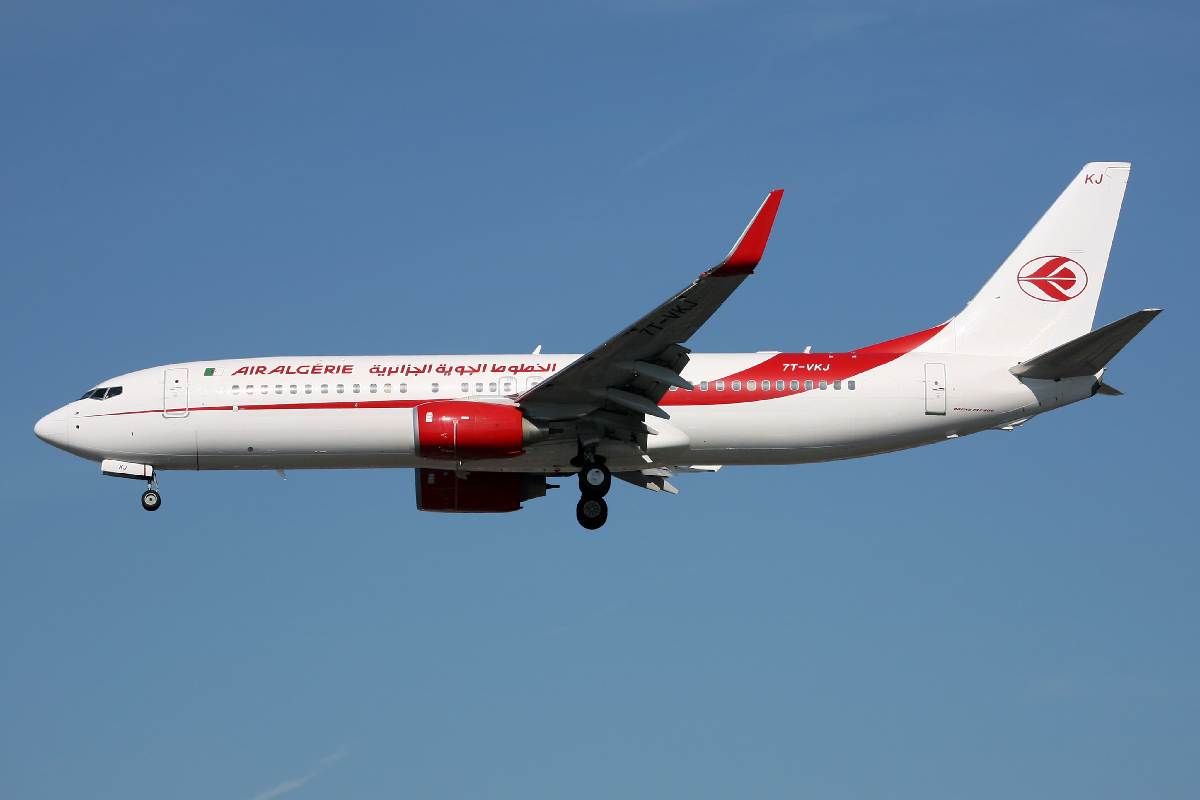 A commercial airplane with Air Algerie livery, featuring red and white design, is mid-flight against a clear blue sky.