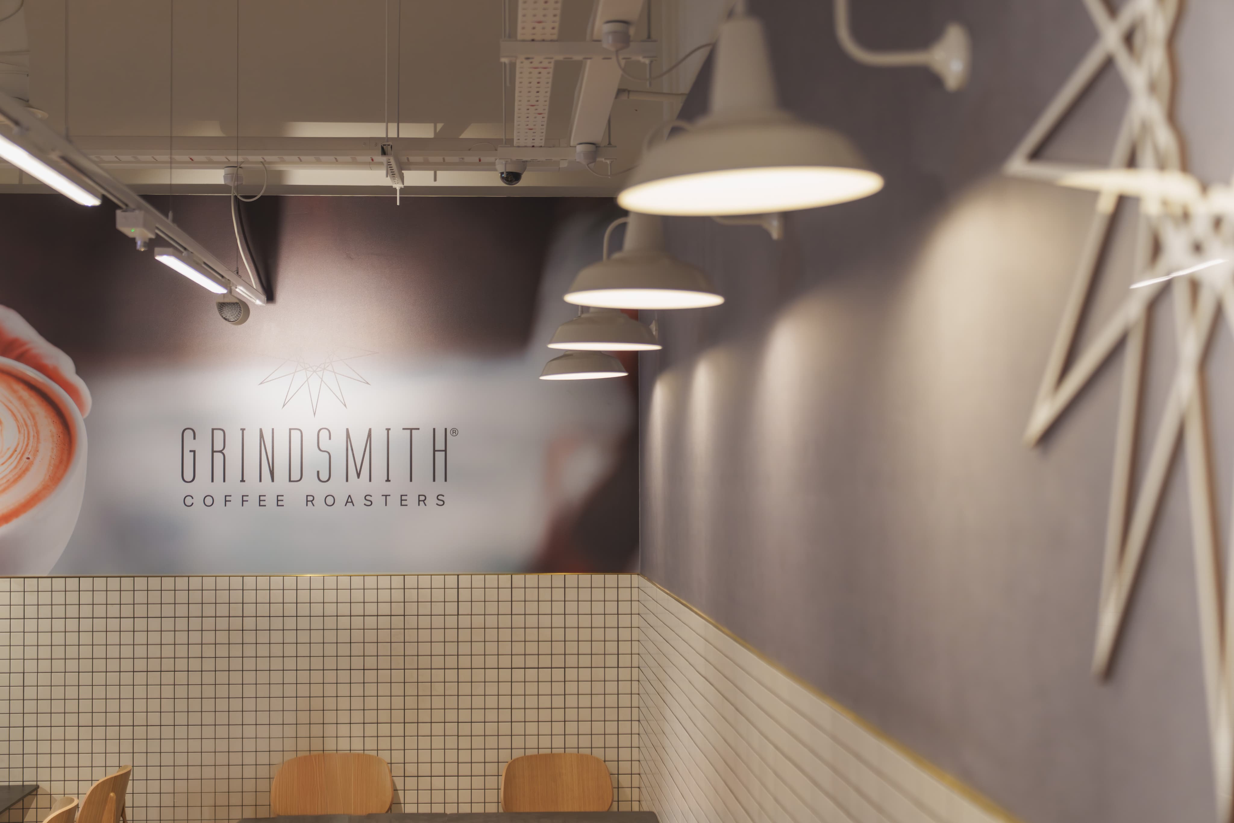 Modern café interior with "Grindsmith Coffee Roasters" sign, tiled walls, and warm pendant lighting above wooden chairs and tables.