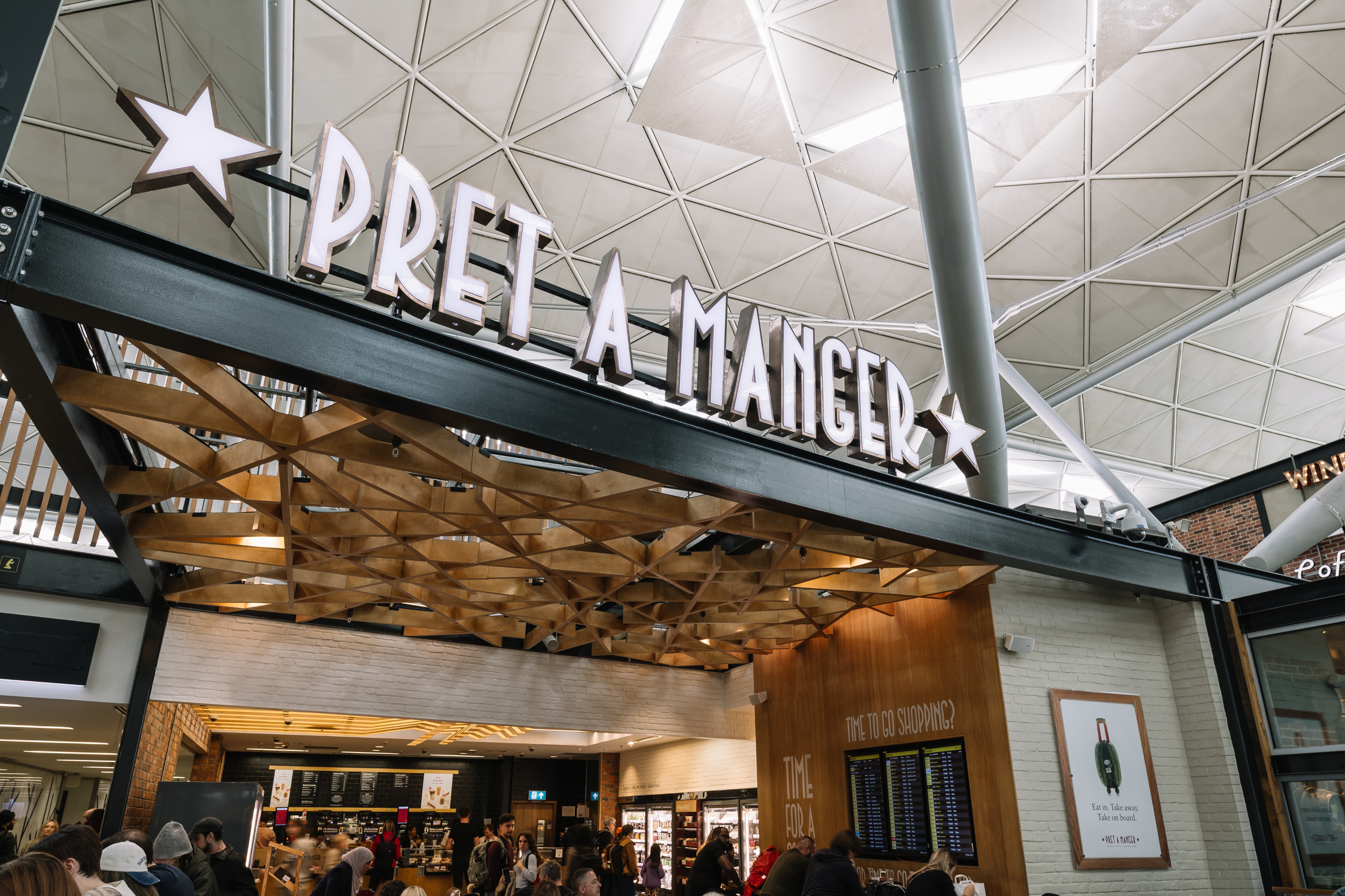Exterior of a Pret A Manger cafe inside a large building with visible wooden roof design and people gathered outside.