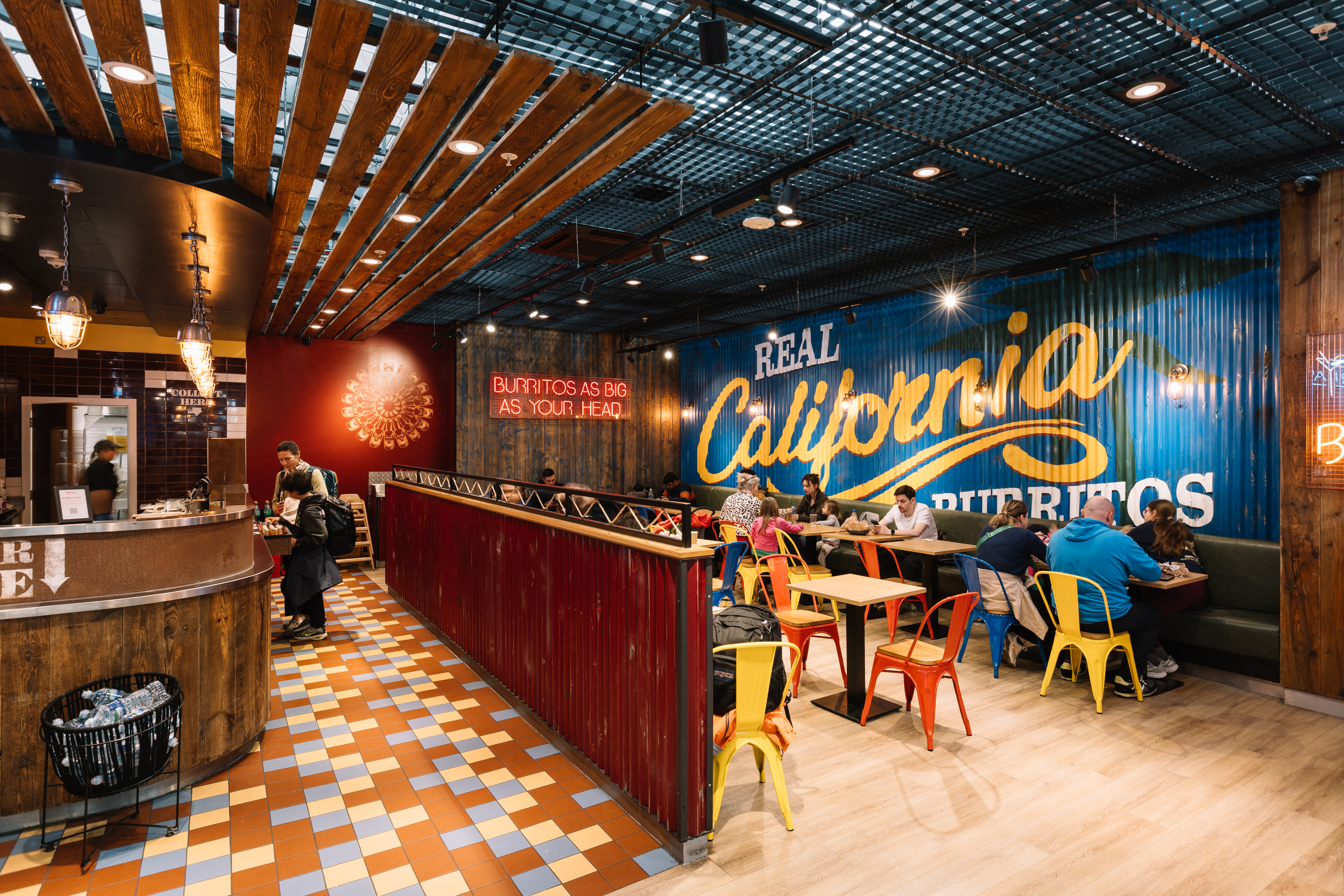 Colourful restaurant with wooden décor, neon signs, and diners seated at tables. Large "Real California Burritos" mural on the wall.