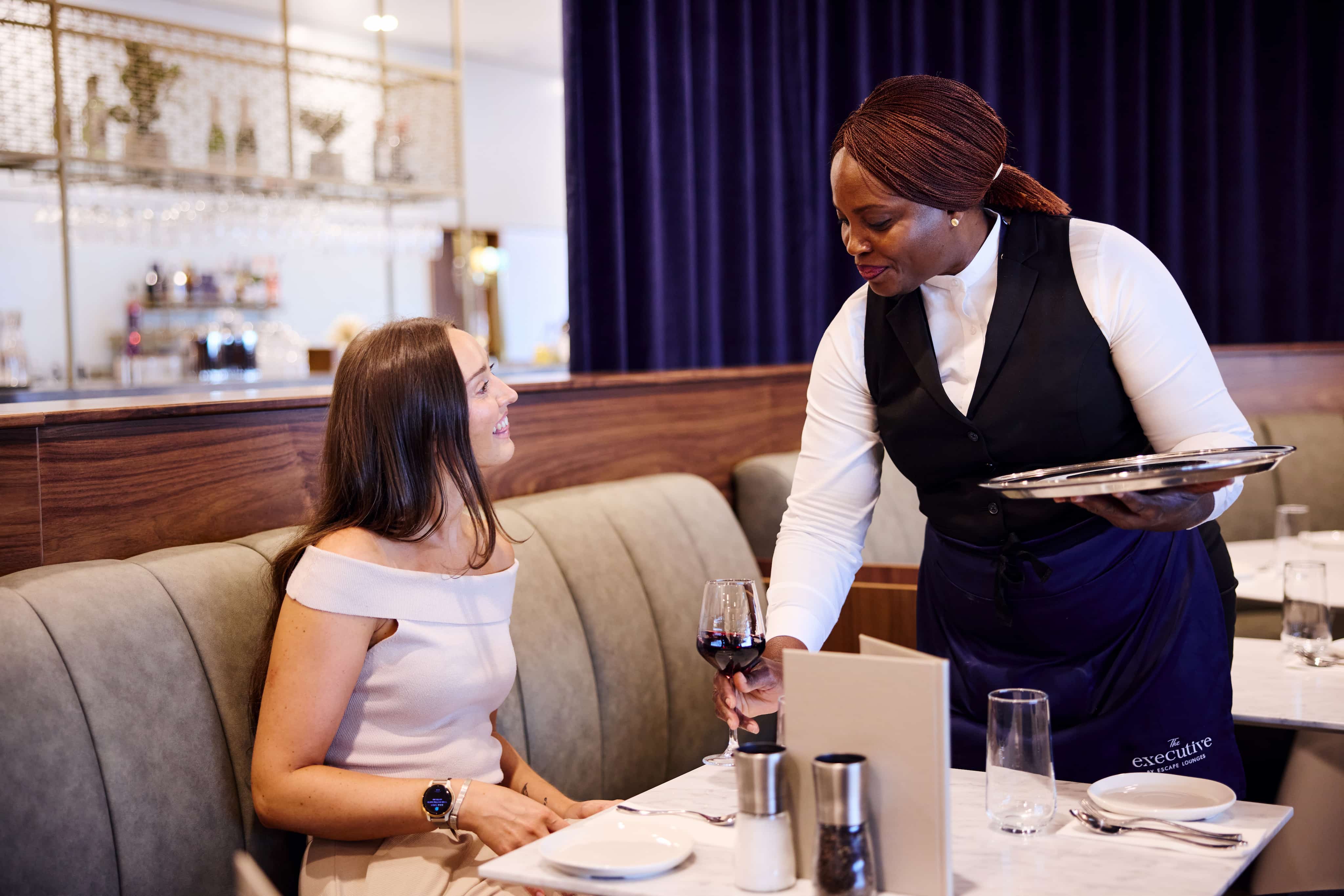 A smiling woman sits at a restaurant table while a server hands her a glass of red wine.