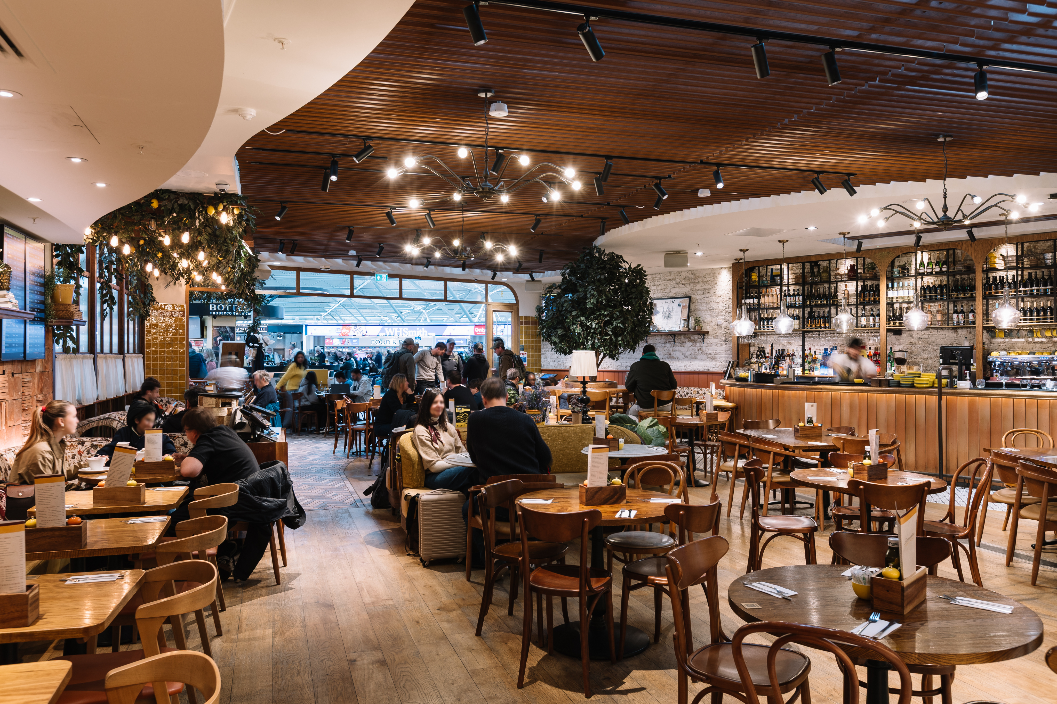 Cosy restaurant interior with warm lighting, wooden tables, passengers dining, and a stocked bar against a backdrop of decorative plants and ambient décor.