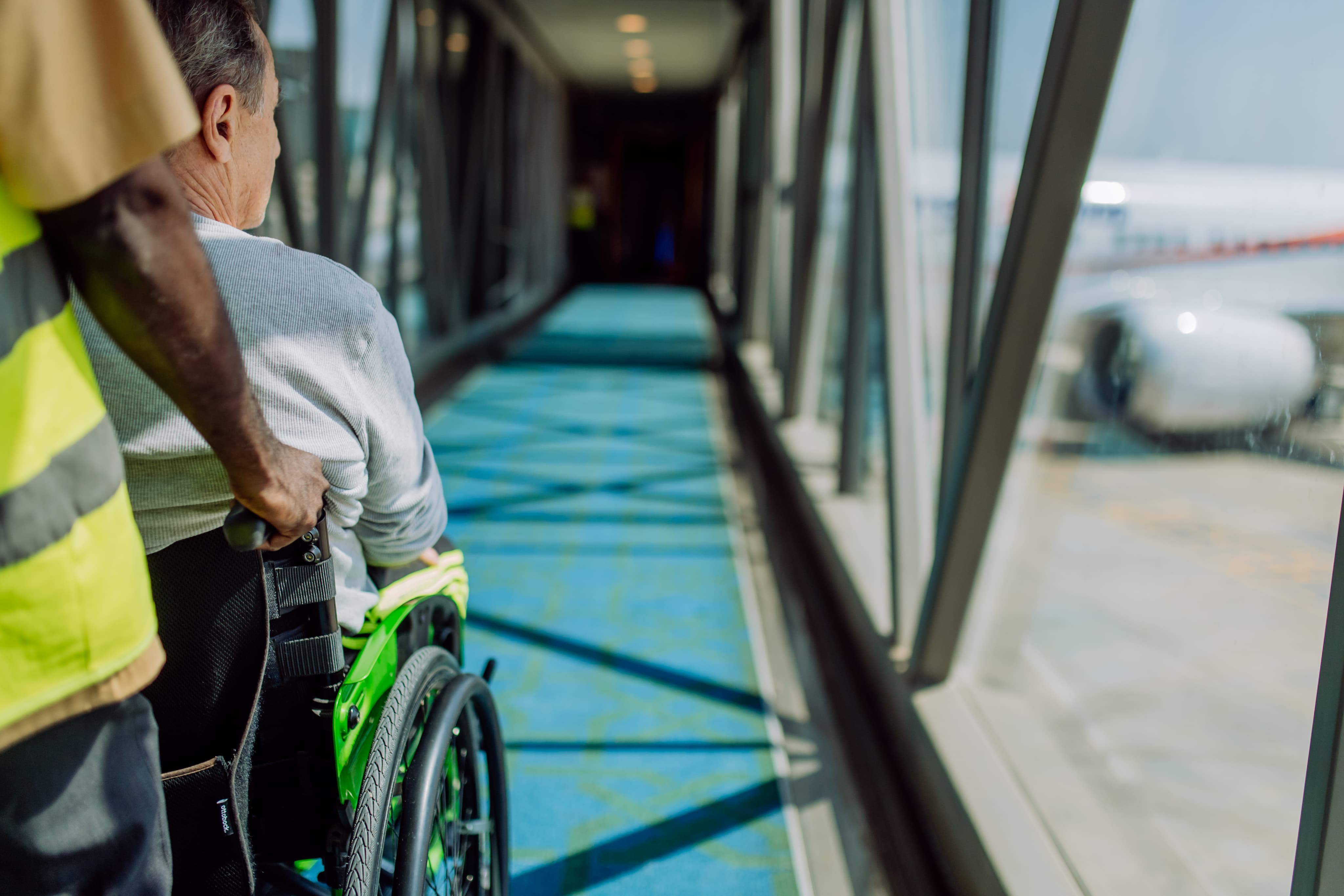 Person in a wheelchair being assisted through an airport jet bridge, with a plane visible outside the window.