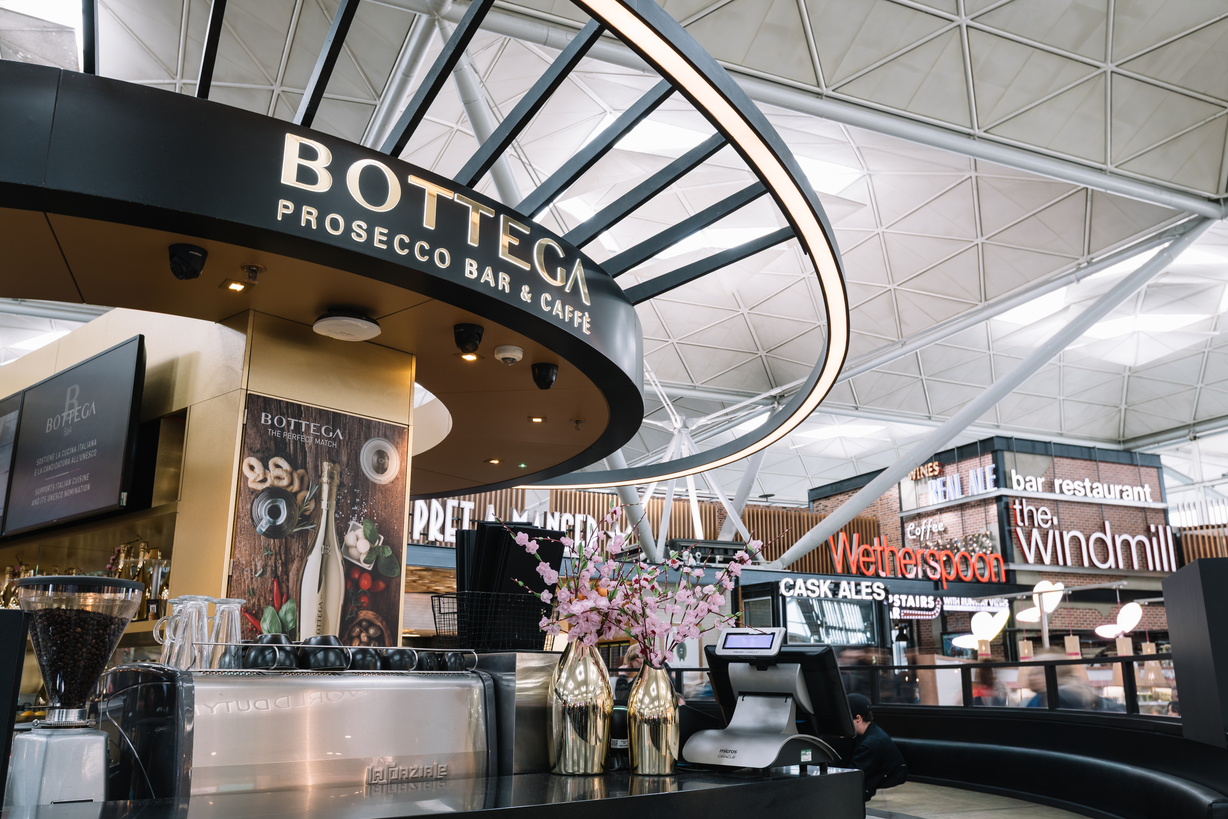 Modern airport café with "Bottega" sign, espresso machine, pink flowers, and seating. Background shows "Wetherspoon" and "The Windmill" signs.