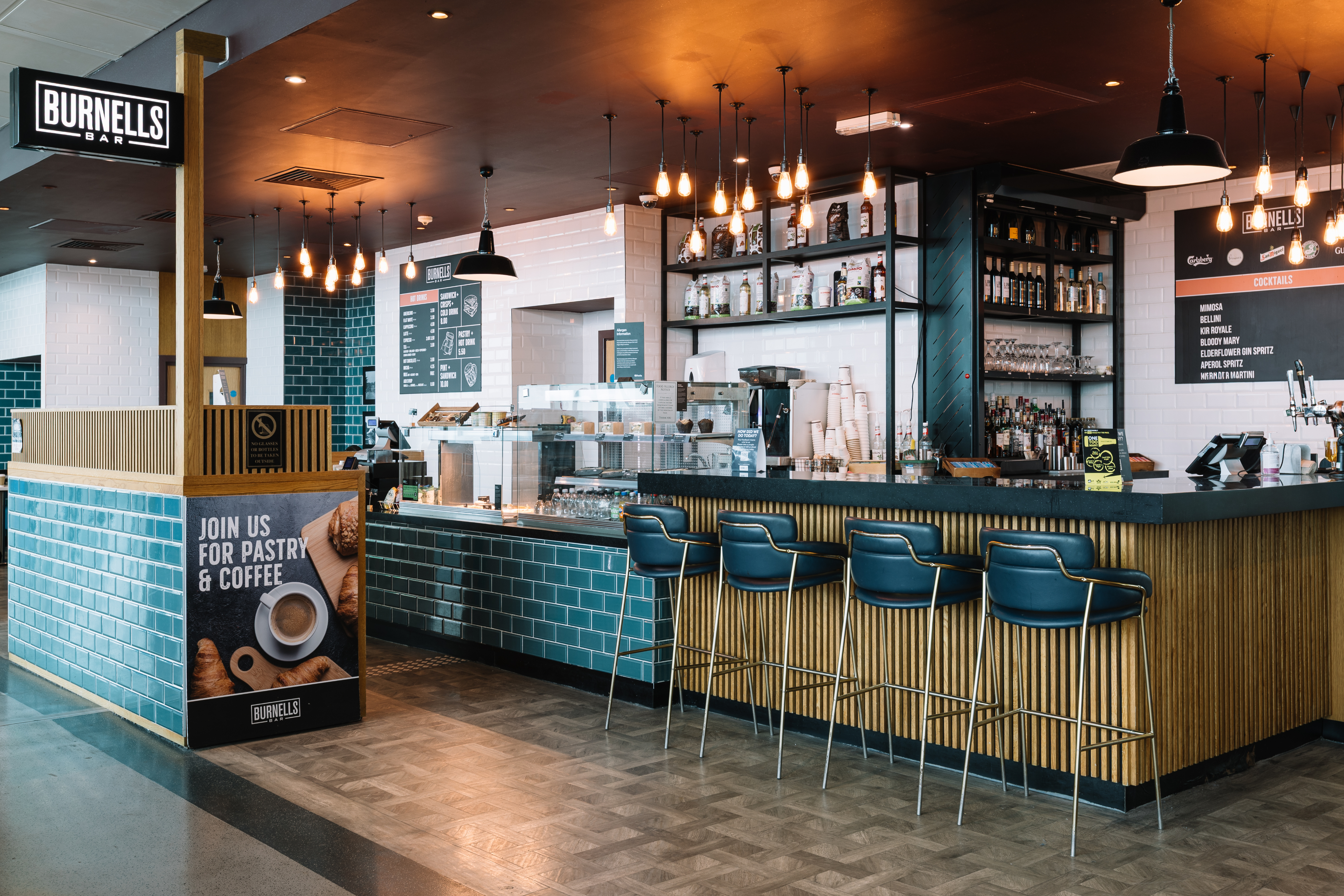 Modern bar with teal tiles, leather barstools, and pendant lighting. Menu boards list coffee and cocktails. Sign promotes pastries and coffee.