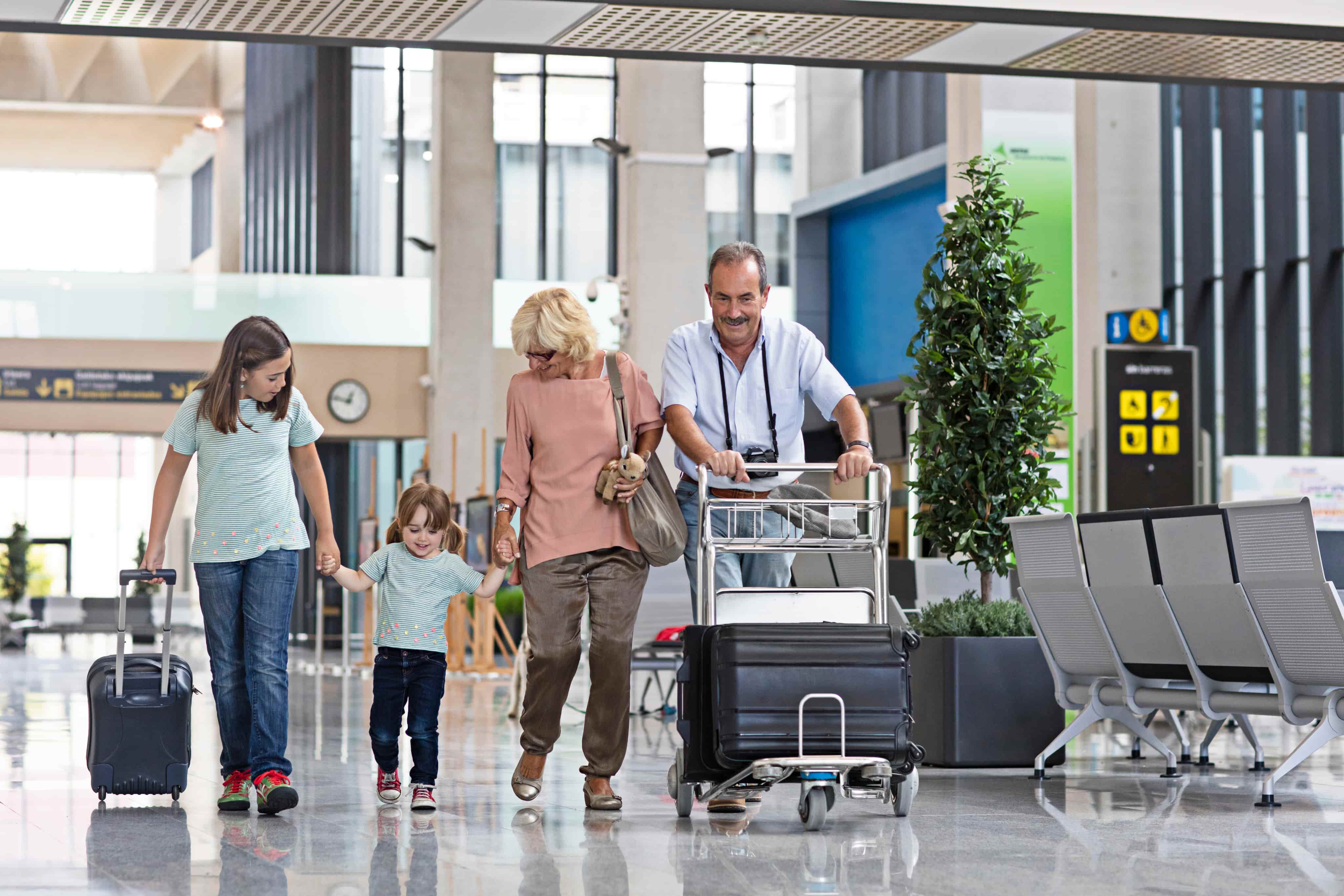 A smiling family with luggage walks through a bright airport terminal, including a man pushing a cart and children pulling suitcases.