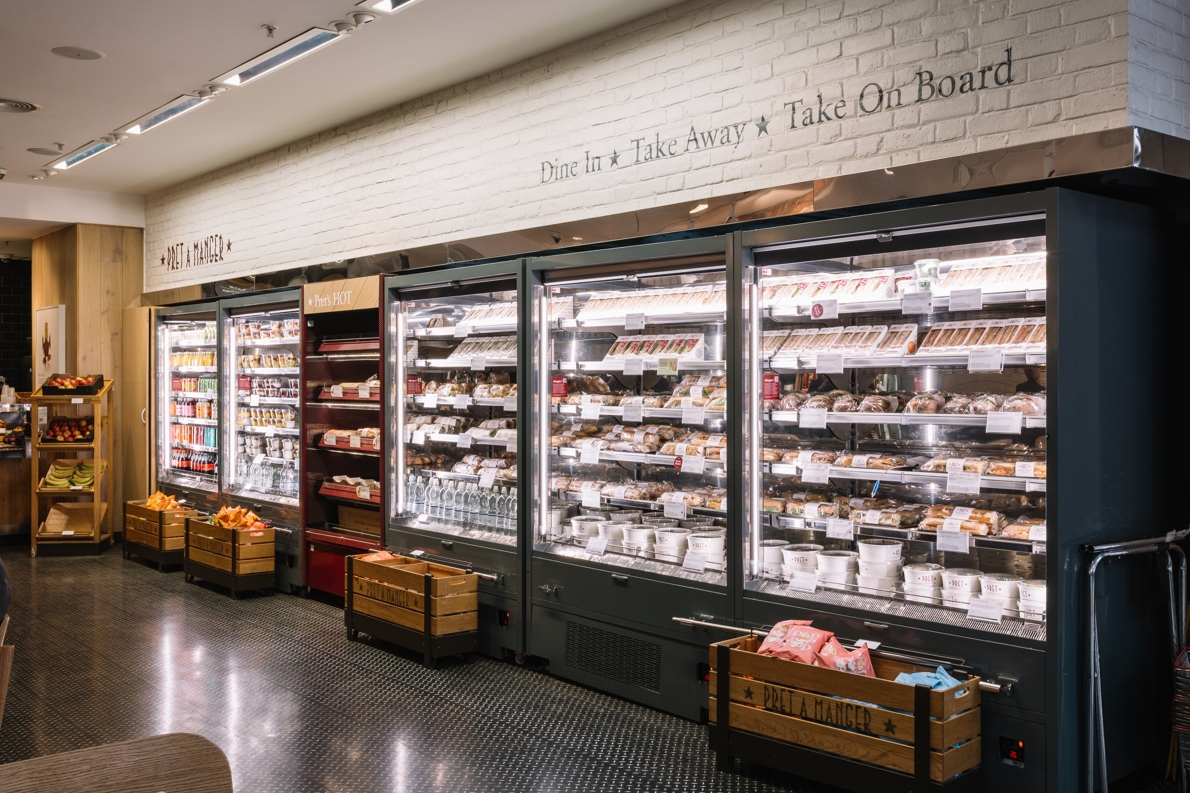A well-lit refrigerated display in a store with sandwiches, drinks, and packaged foods. Wooden crates hold fresh fruit and snacks.