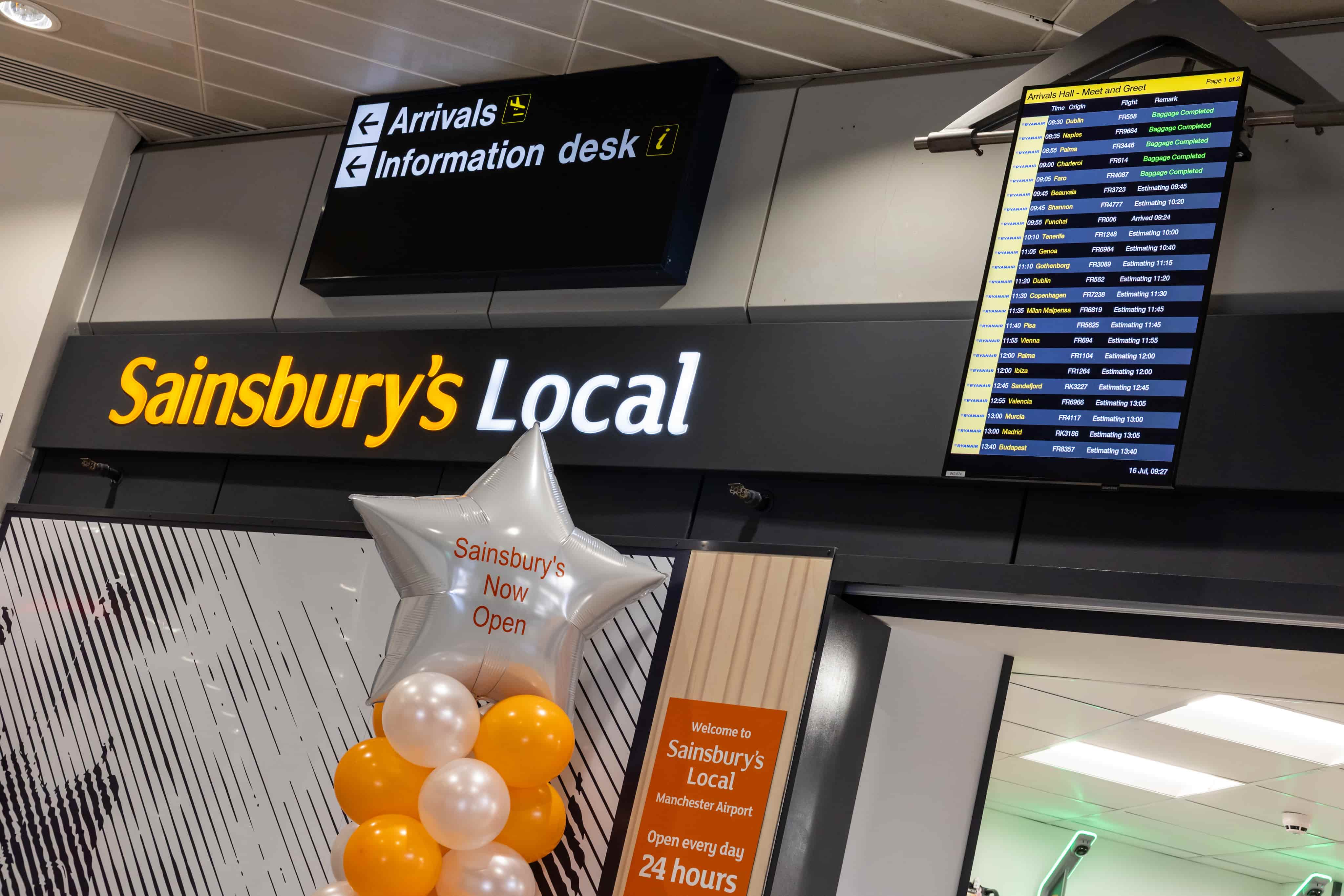 Sainsbury's Local store at Manchester Airport with "Now Open" balloons. Flight information screen and ceiling signs visible.