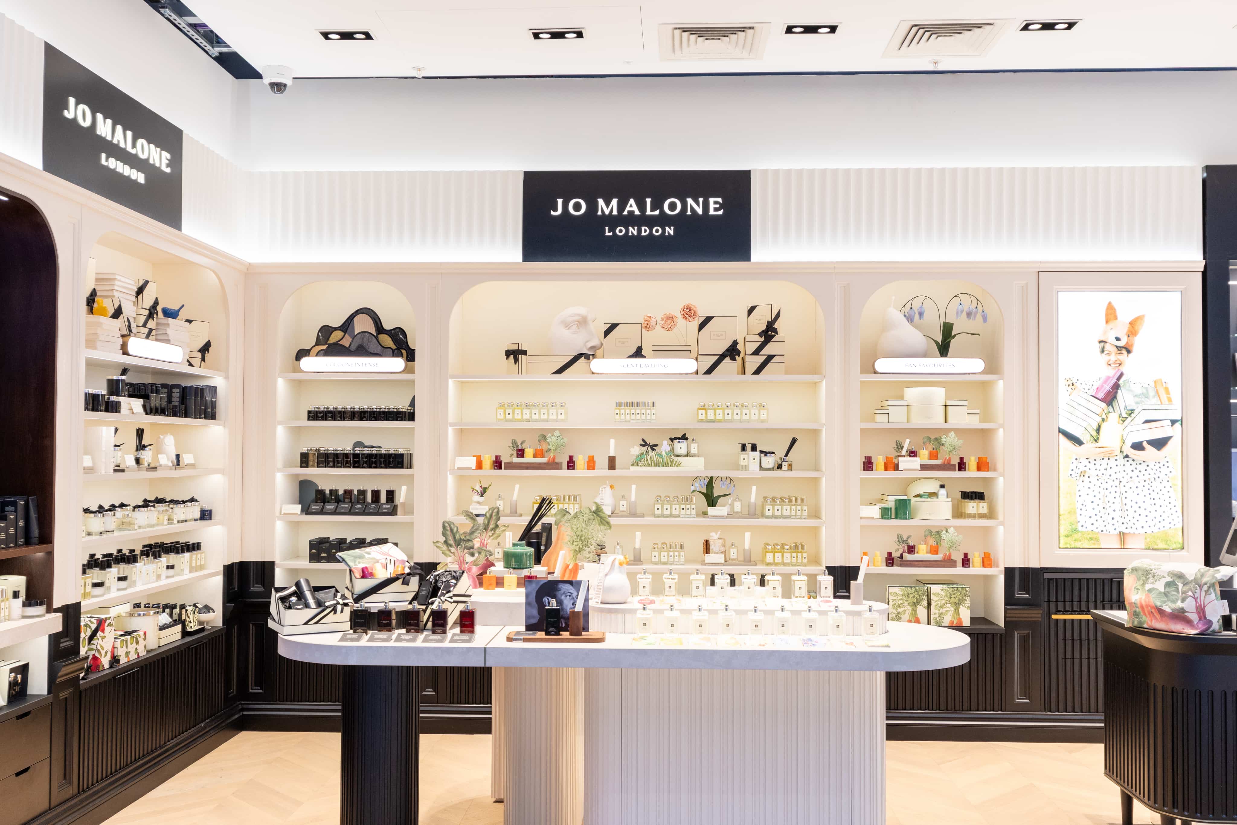 A Jo Malone London store interior with shelves displaying perfumes and home fragrances, a central counter with products, and elegant decor.