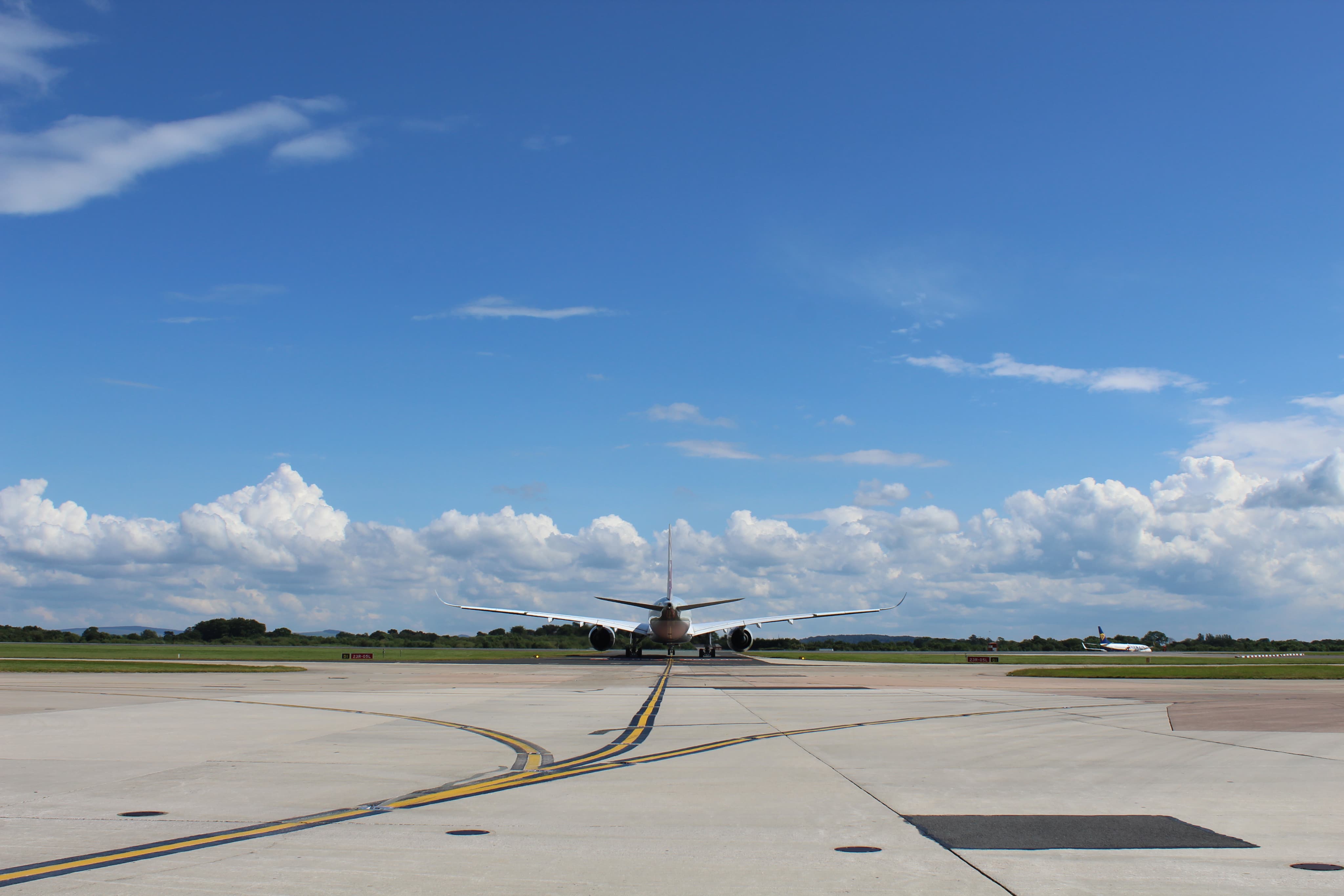 Airplane taxiing on a runway under a clear blue sky filled with scattered clouds, with grassy fields in the background.