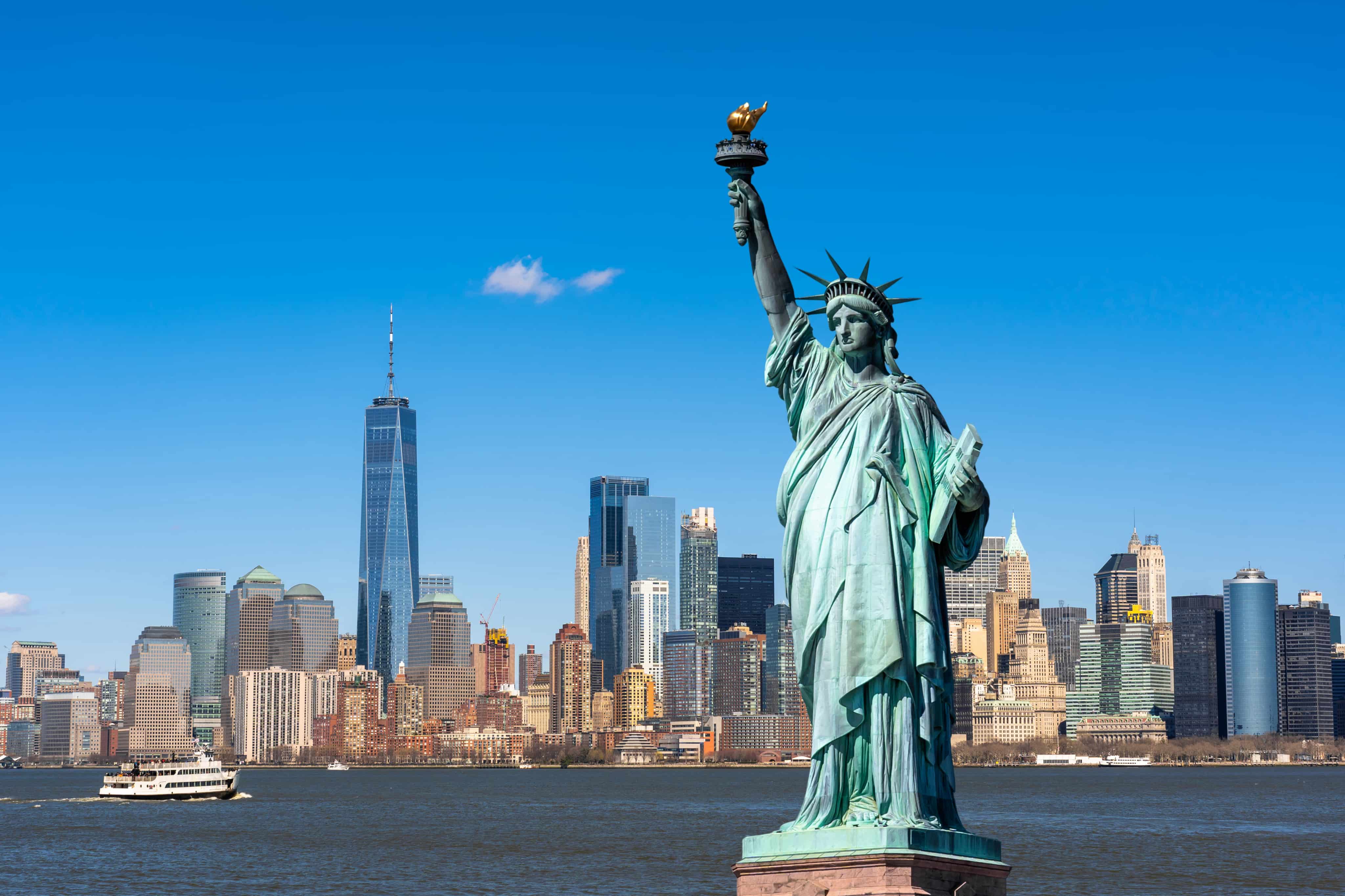 Statue of Liberty with torch raised, overlooking New York City skyline and Hudson River, under a clear blue sky.