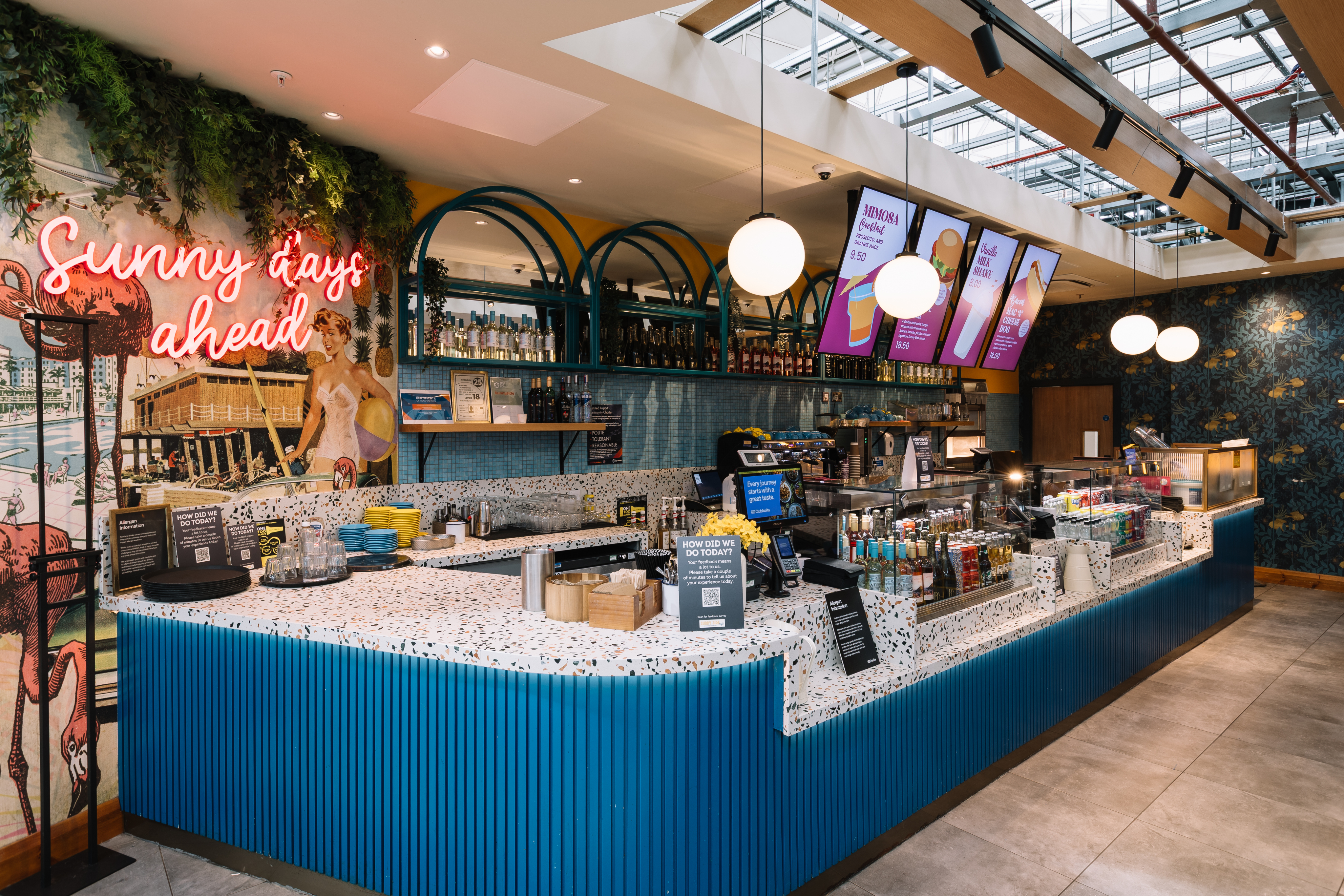 A modern café with a colourful counter and neon sign reading "Sunny days ahead." Hanging lights, menu screens, and tropical-themed decor are visible.