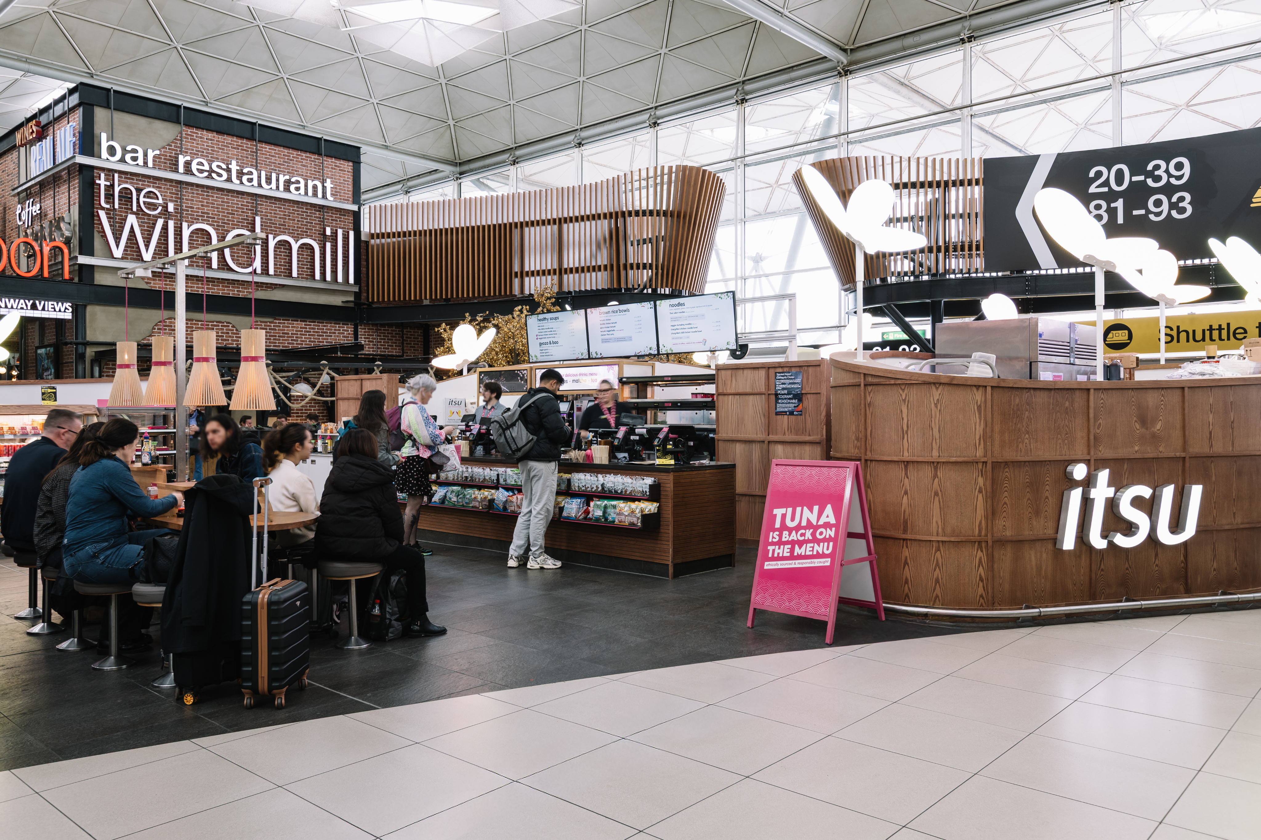 People dining and ordering at a modern airport food court featuring "itsu" and "the windmill" eateries. A sign highlights tuna on the menu.