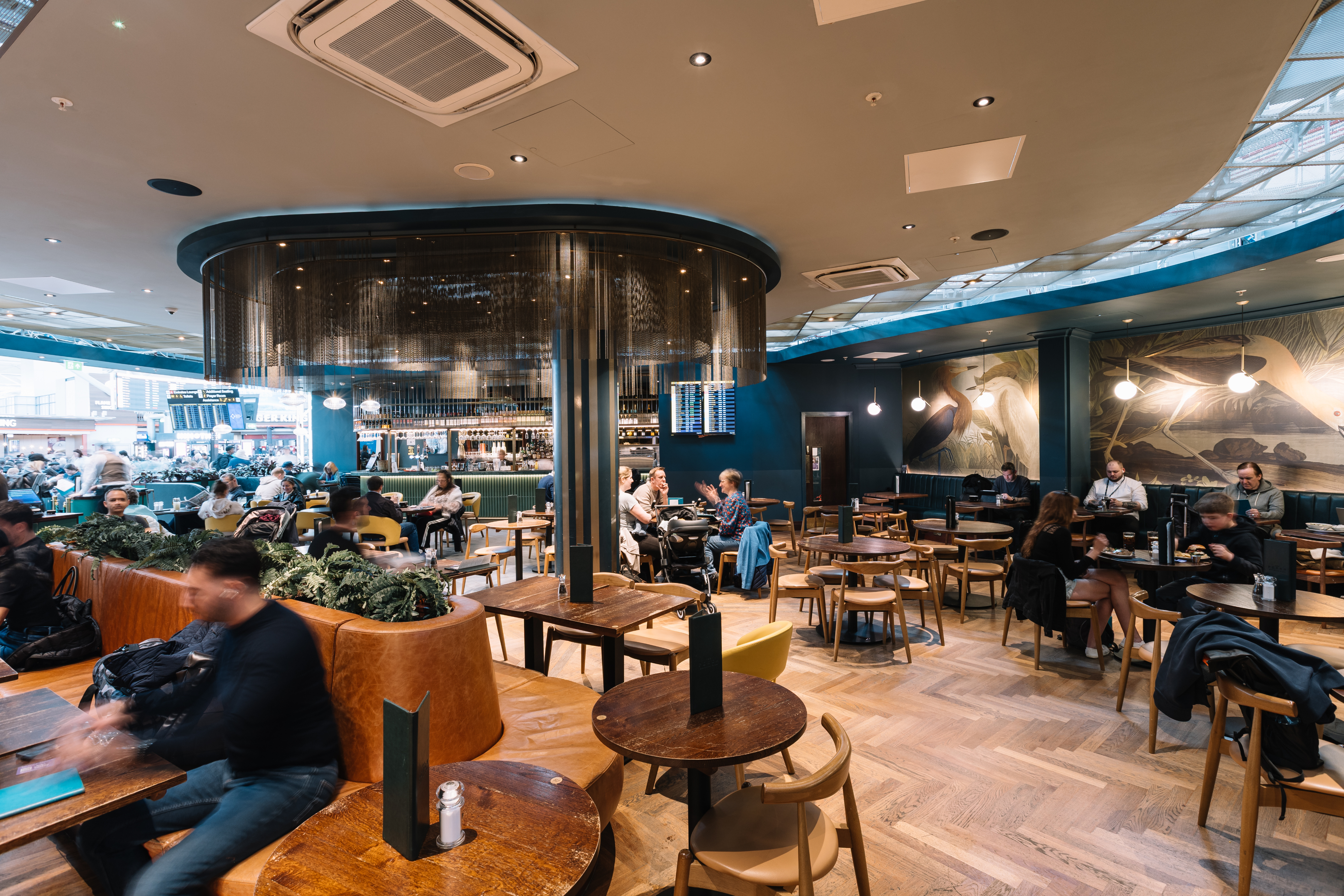 Spacious café with modern décor, wooden tables, and customers dining and socialising. Wall art depicts birds.
