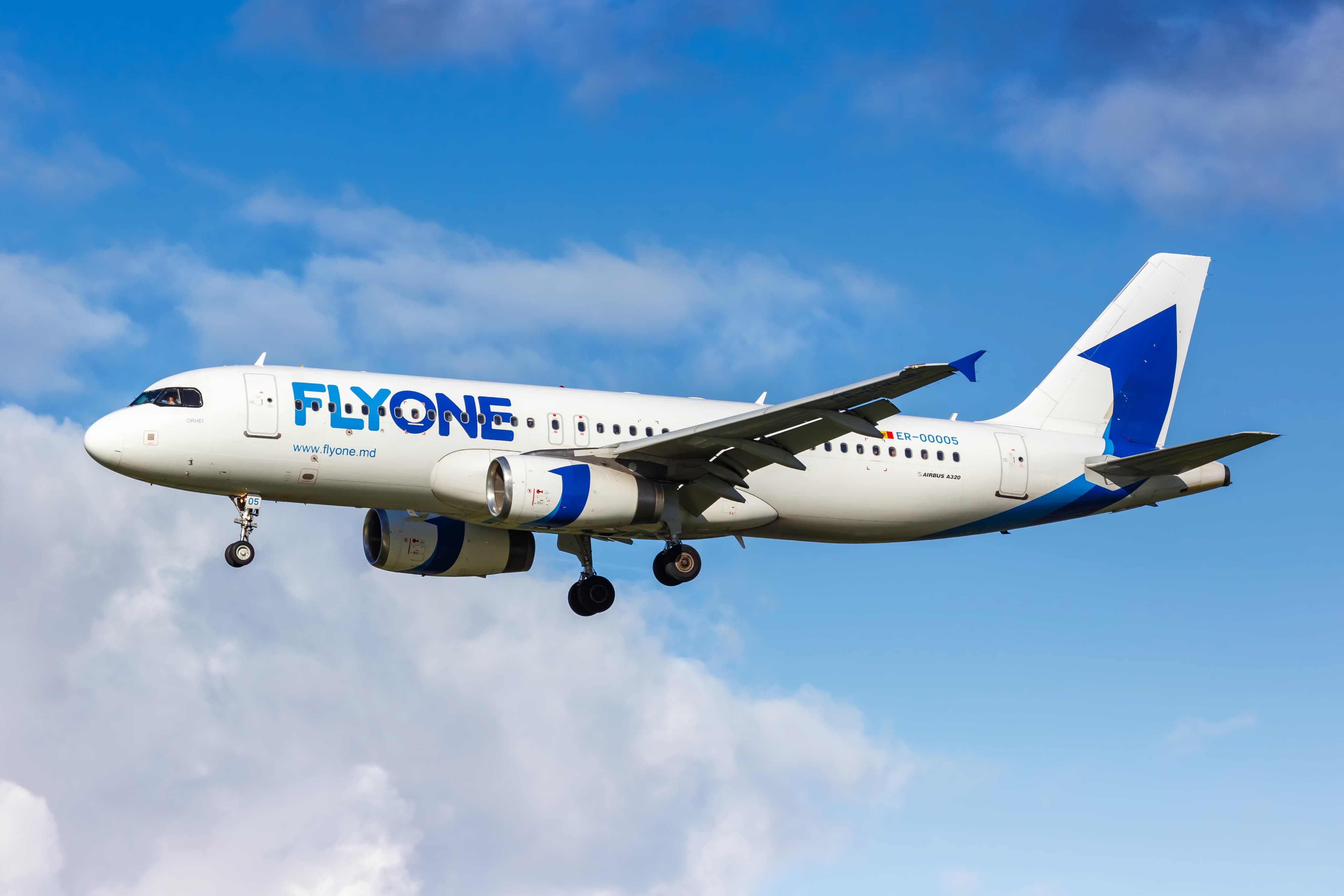 White and blue FlyOne airplane flying against a backdrop of blue sky and clouds.