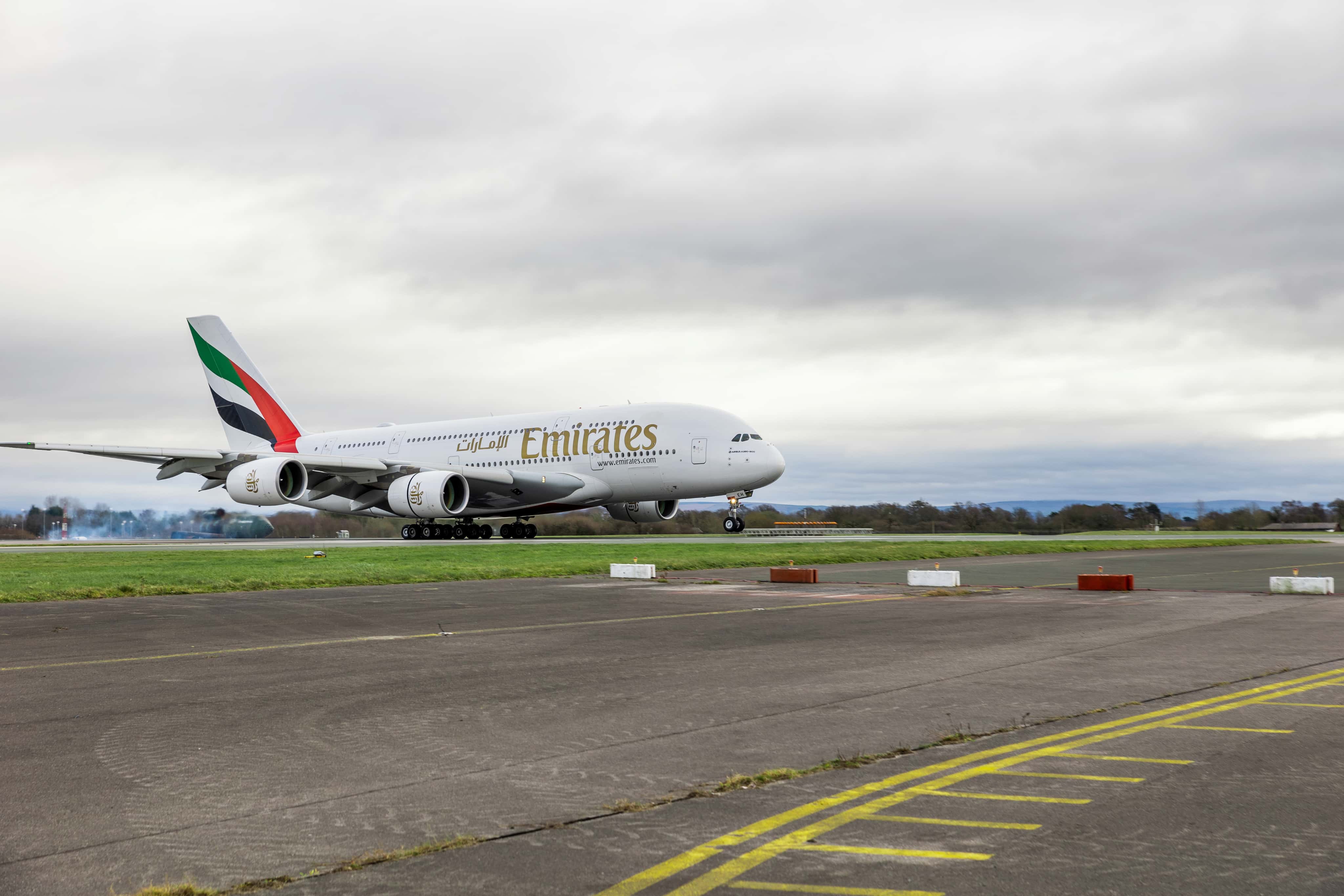 An Emirates Airbus A380 lands on a runway surrounded by grass, under a cloudy sky.