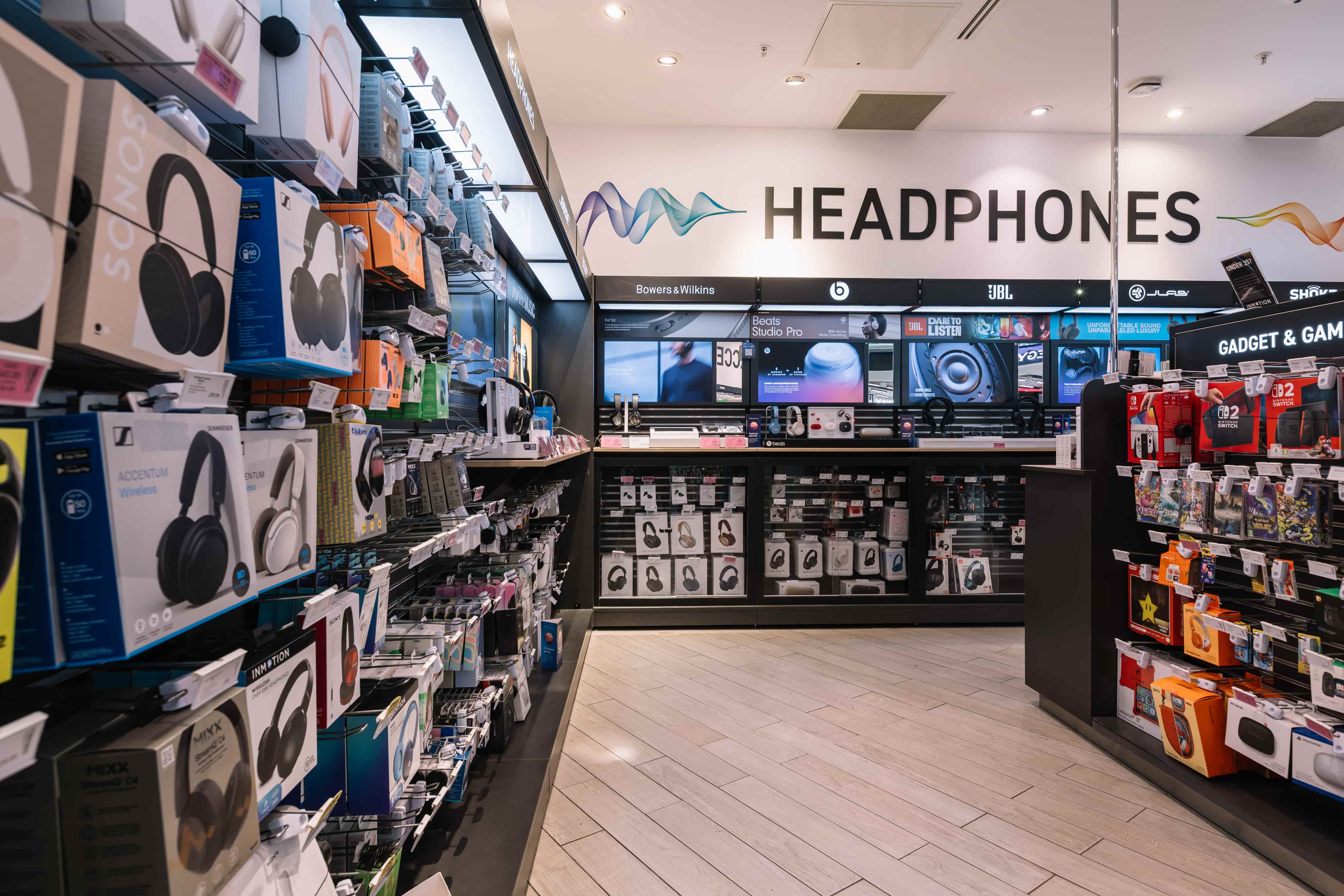 Electronics store headphone section, displaying various branded headphones and accessories in colourful packaging on shelves.