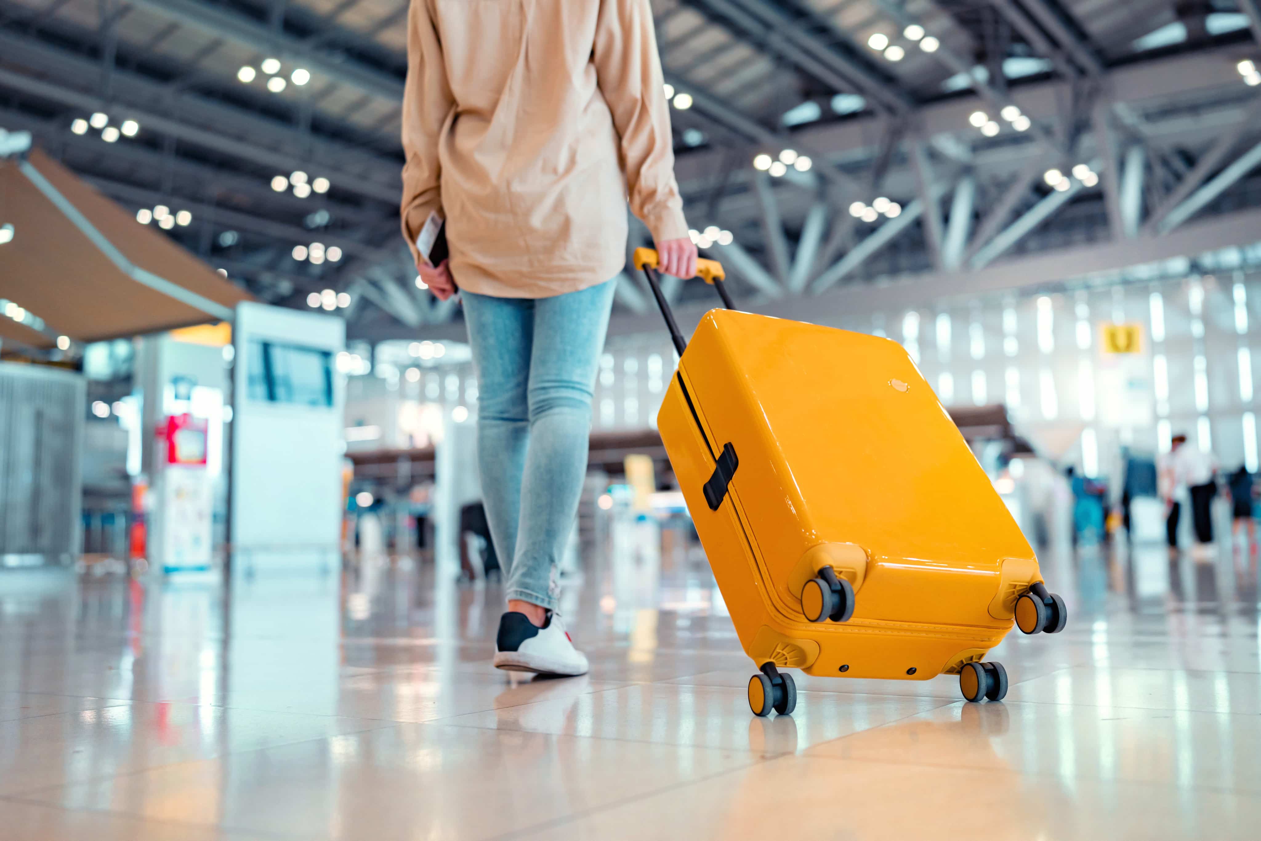 Person walking through an airport, pulling a bright yellow suitcase with four wheels, wearing blue jeans and white sneakers.