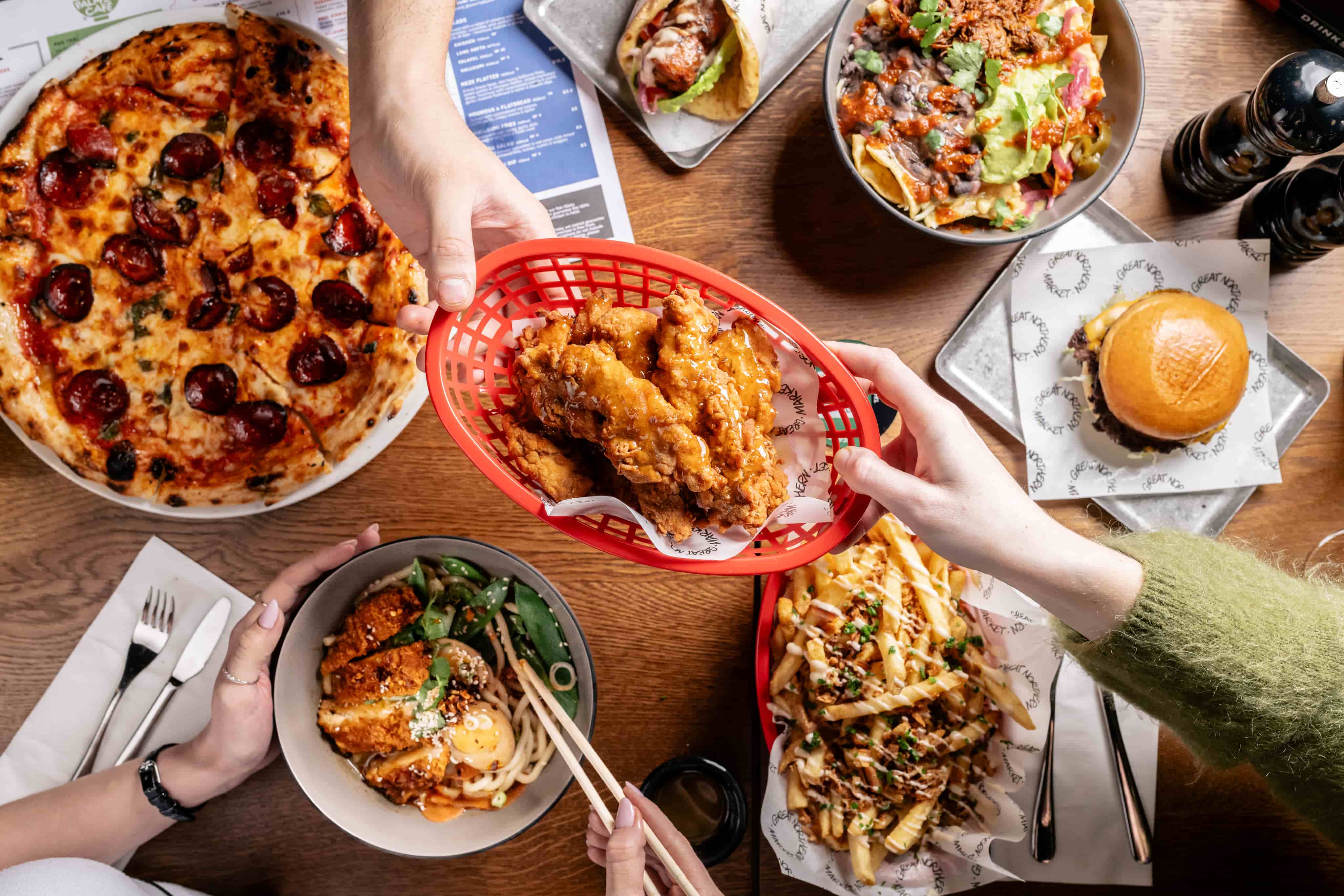 A table with pizza, fried chicken, nachos, fries, ramen, and a burger, with hands sharing the food.