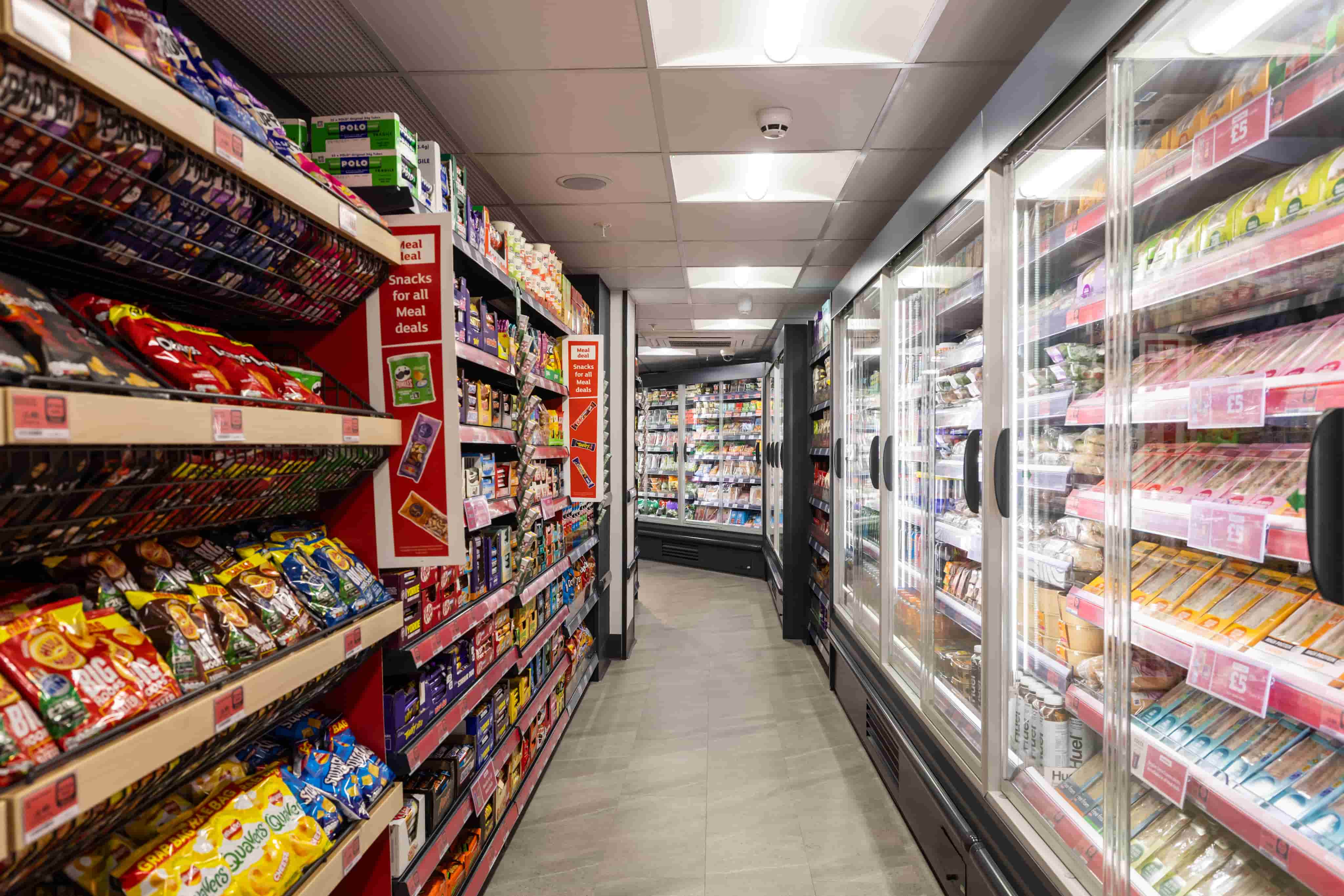 Grocery store aisle with shelves of snacks on the left and refrigerated food items on the right, under bright overhead lights.