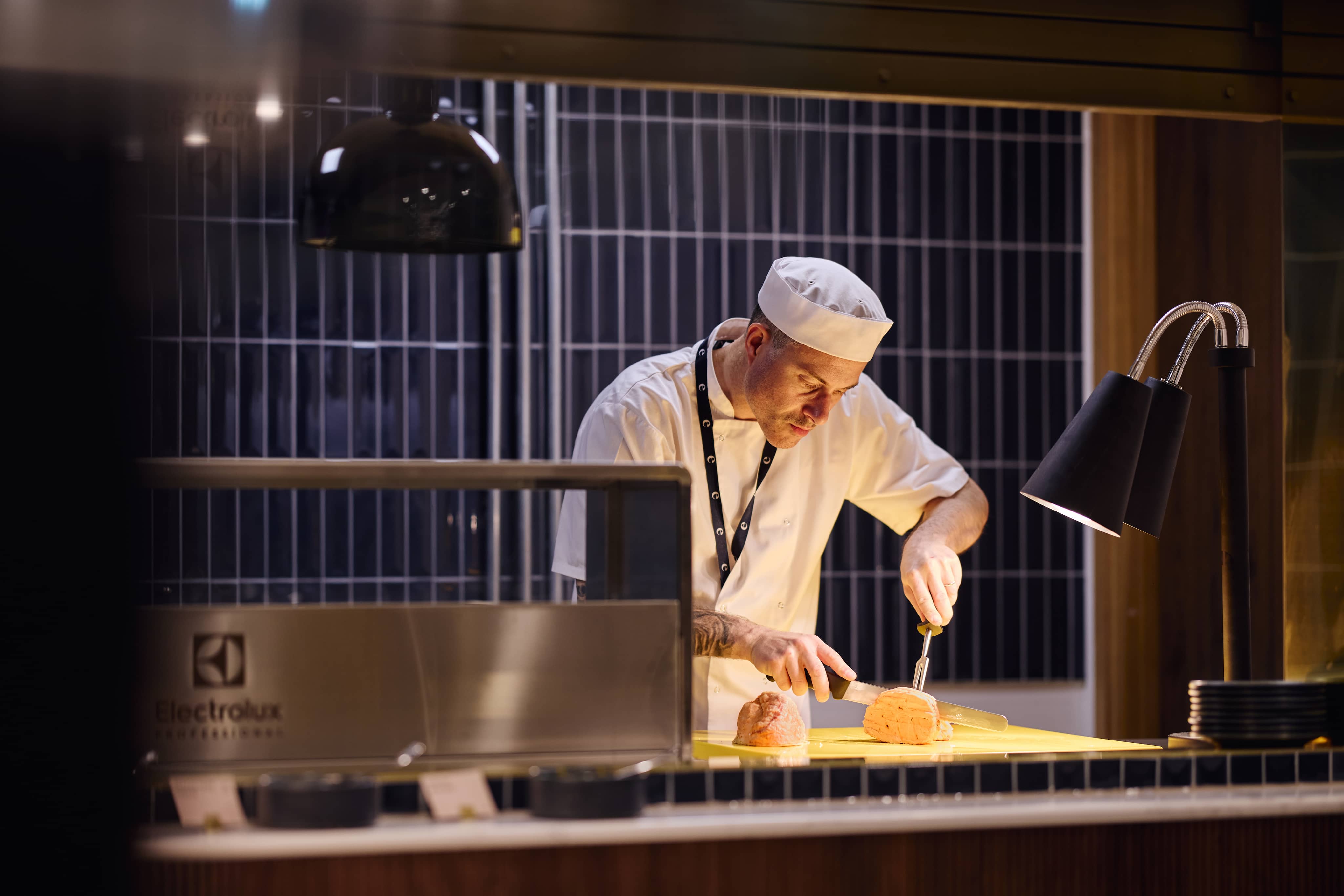 A chef in a white uniform slices meat under warm lighting in a professional kitchen setting.
