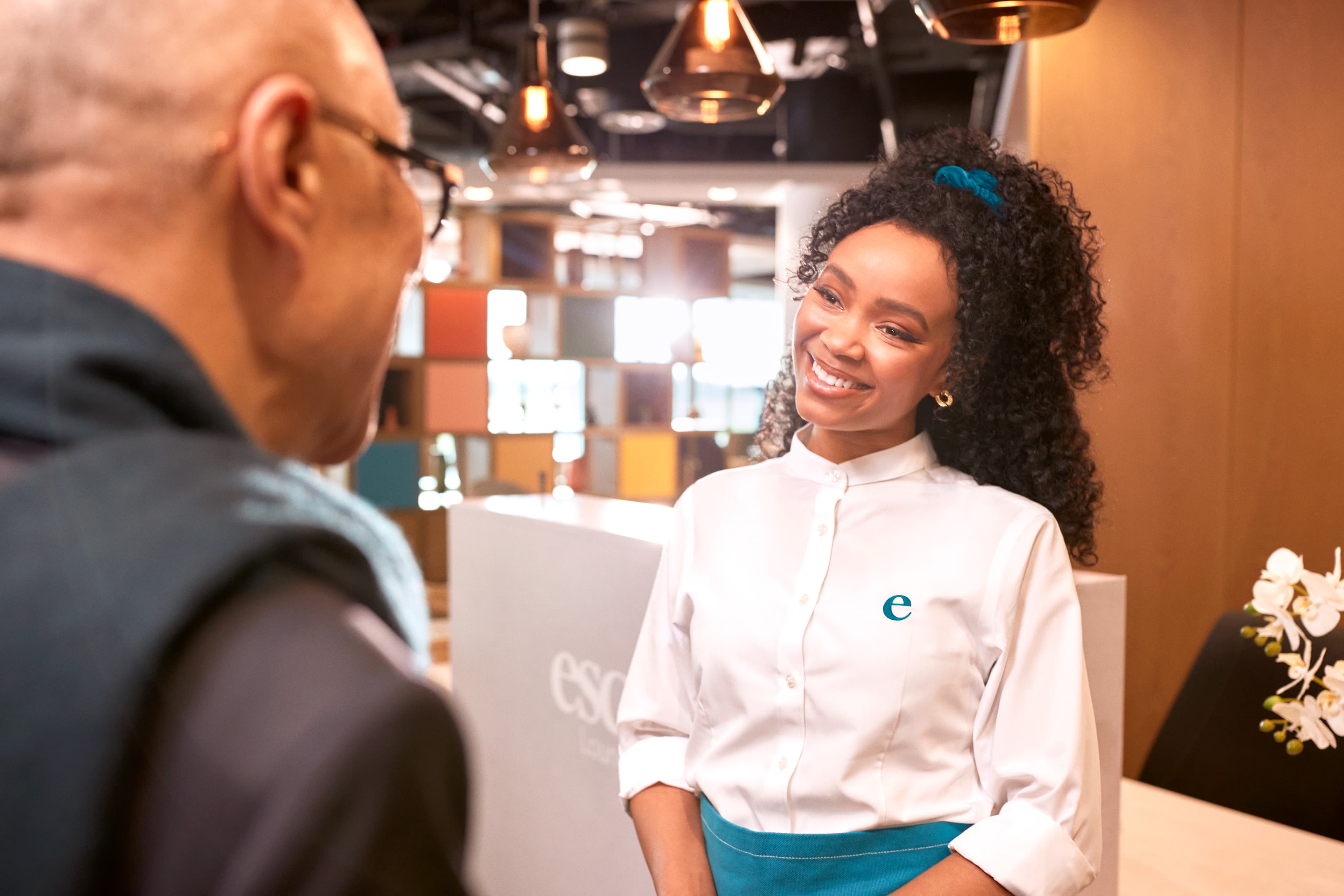 A friendly woman in a uniform with an "e" logo smiles while interacting with a customer at a modern reception area.