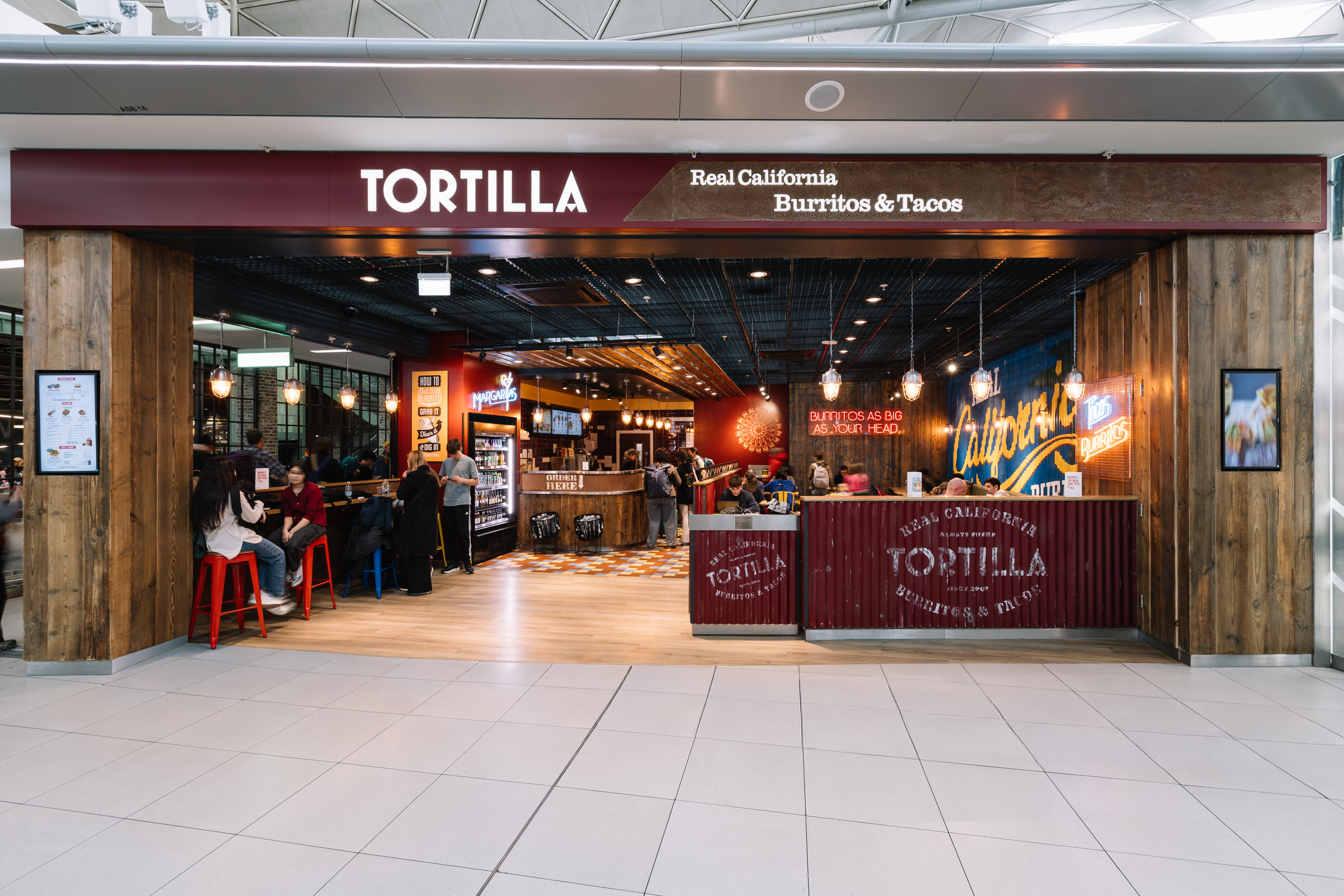 Restaurant entrance labelled "Tortilla" with seating area and counter, offering burritos and tacos under warm hanging lights.