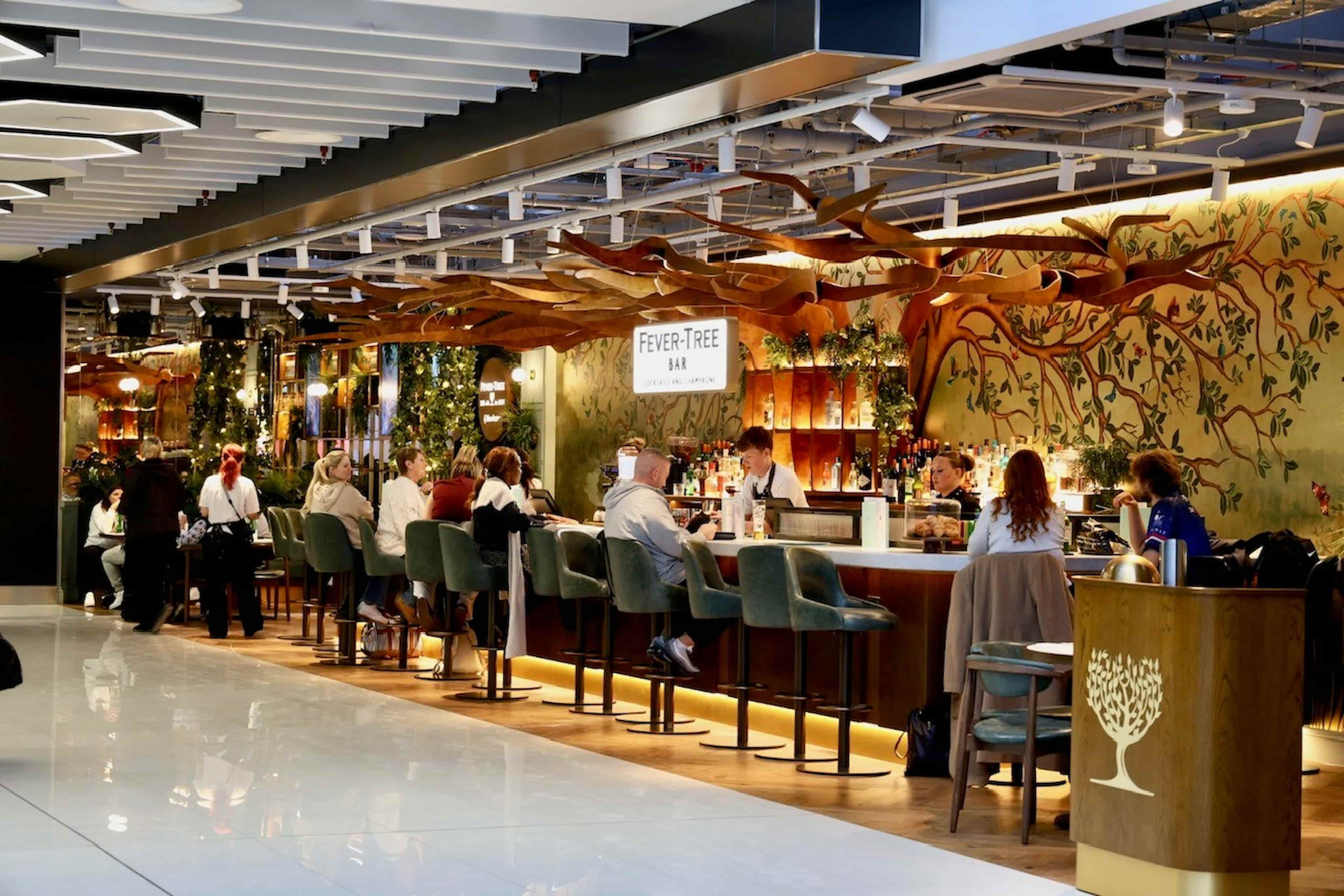 A modern bar with people seated, featuring a wooden counter, green upholstered chairs, and leafy decor on the walls and ceiling.