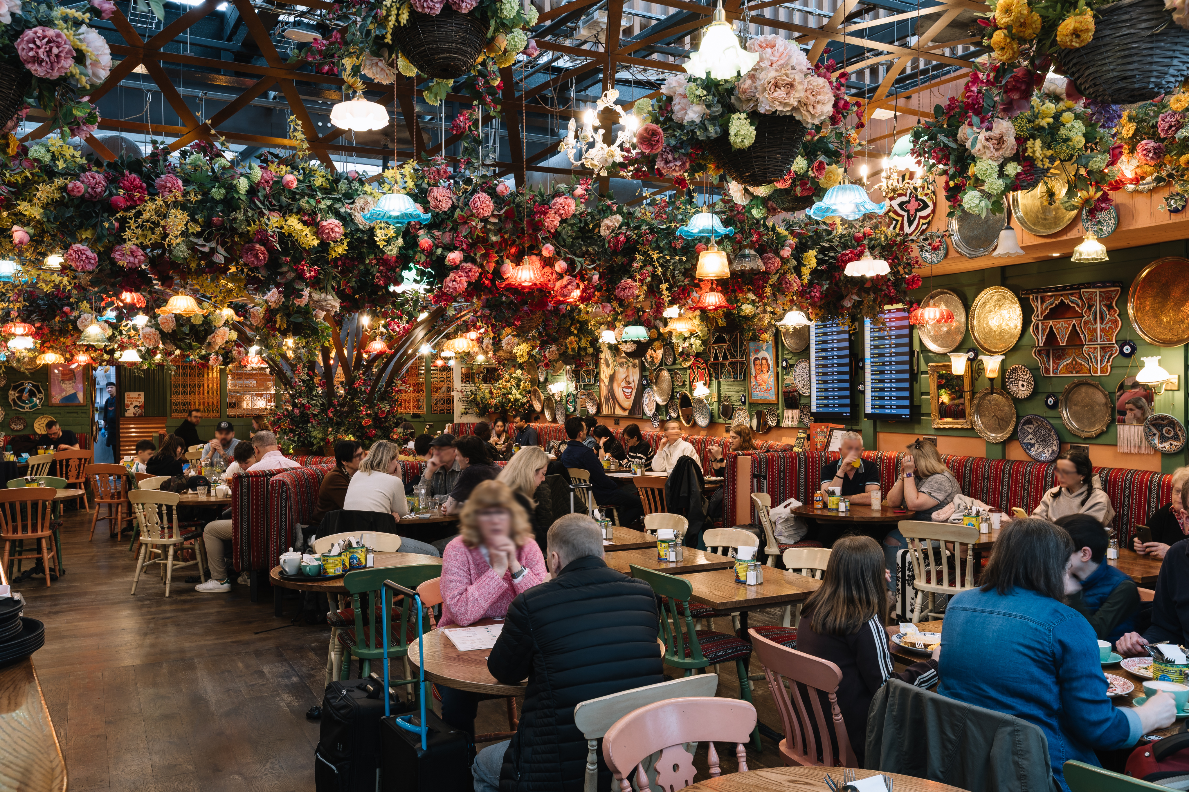 Vibrant restaurant interior with floral décor, colourful hanging lamps, and patrons seated at wooden tables, creating a lively atmosphere.