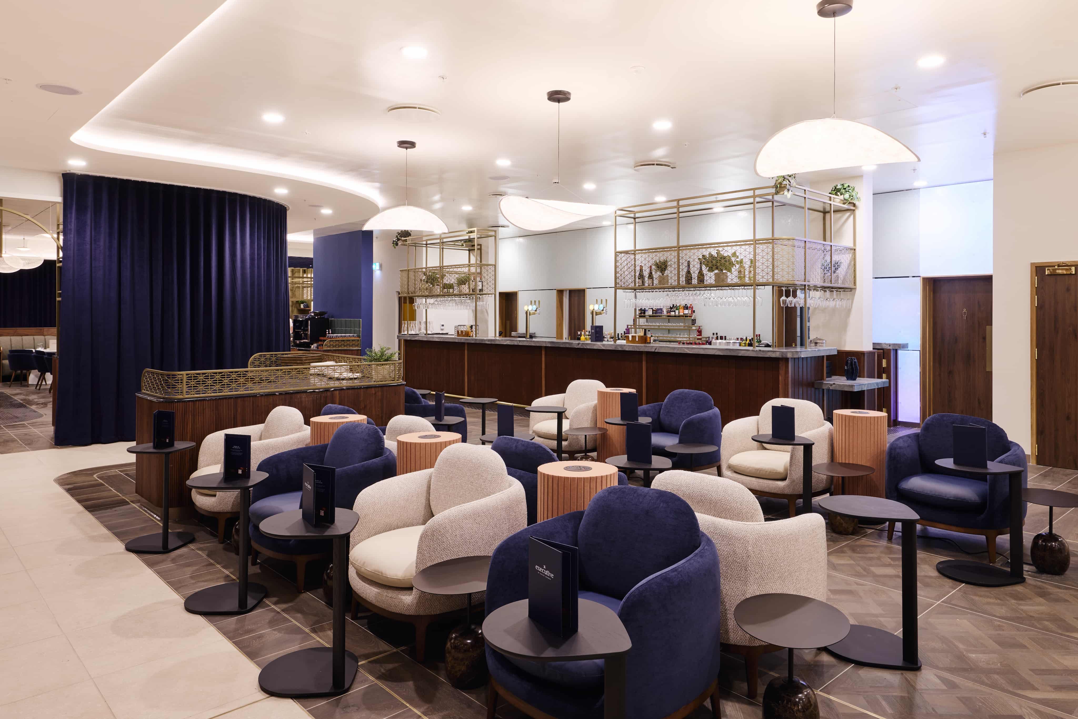 Elegant lounge with plush blue and beige chairs, small round tables, and a bar in the background, under soft, modern lighting.