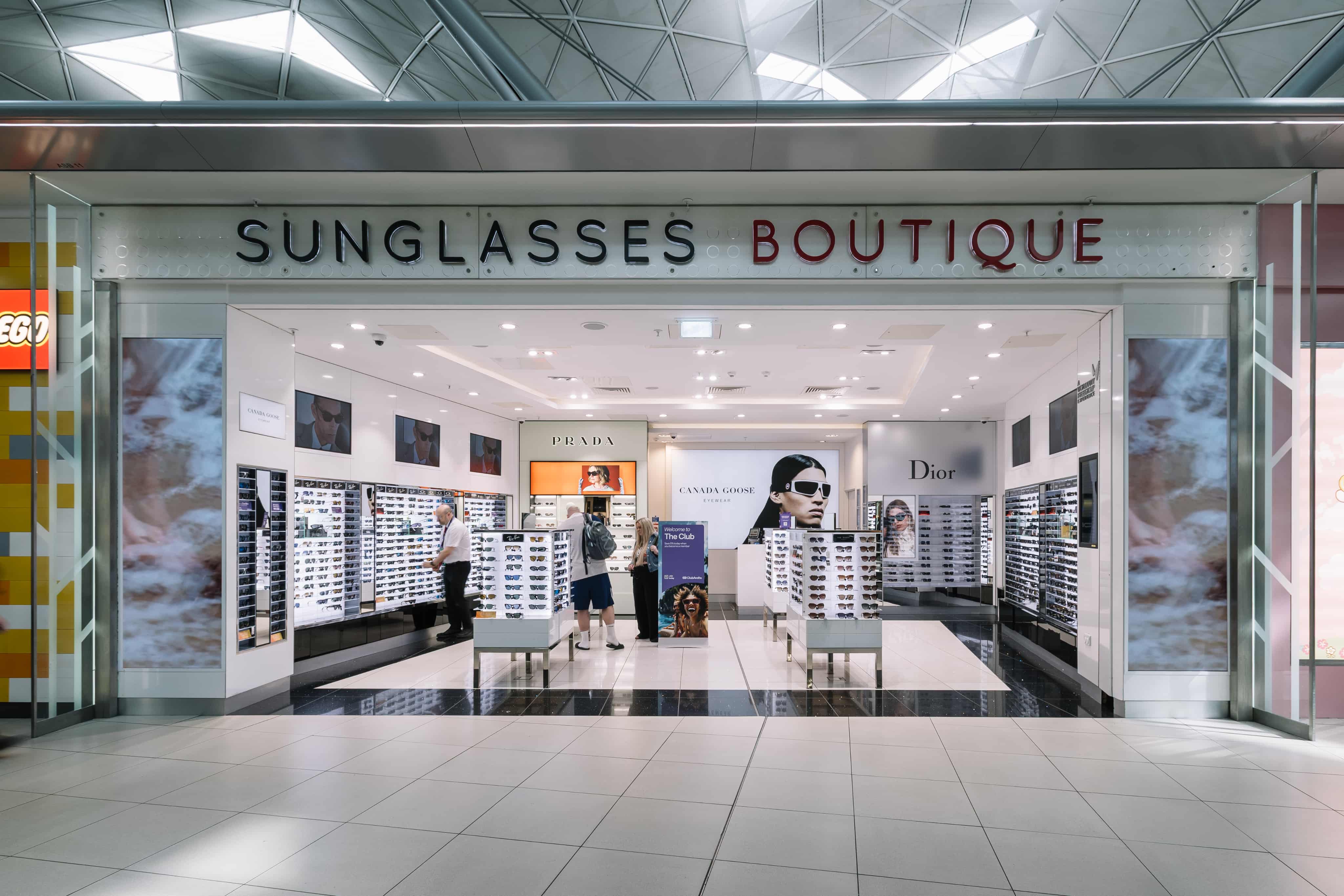 Storefront of "Sunglasses Boutique" with displays of eyewear from brands like Prada and Dior, and customers browsing inside.