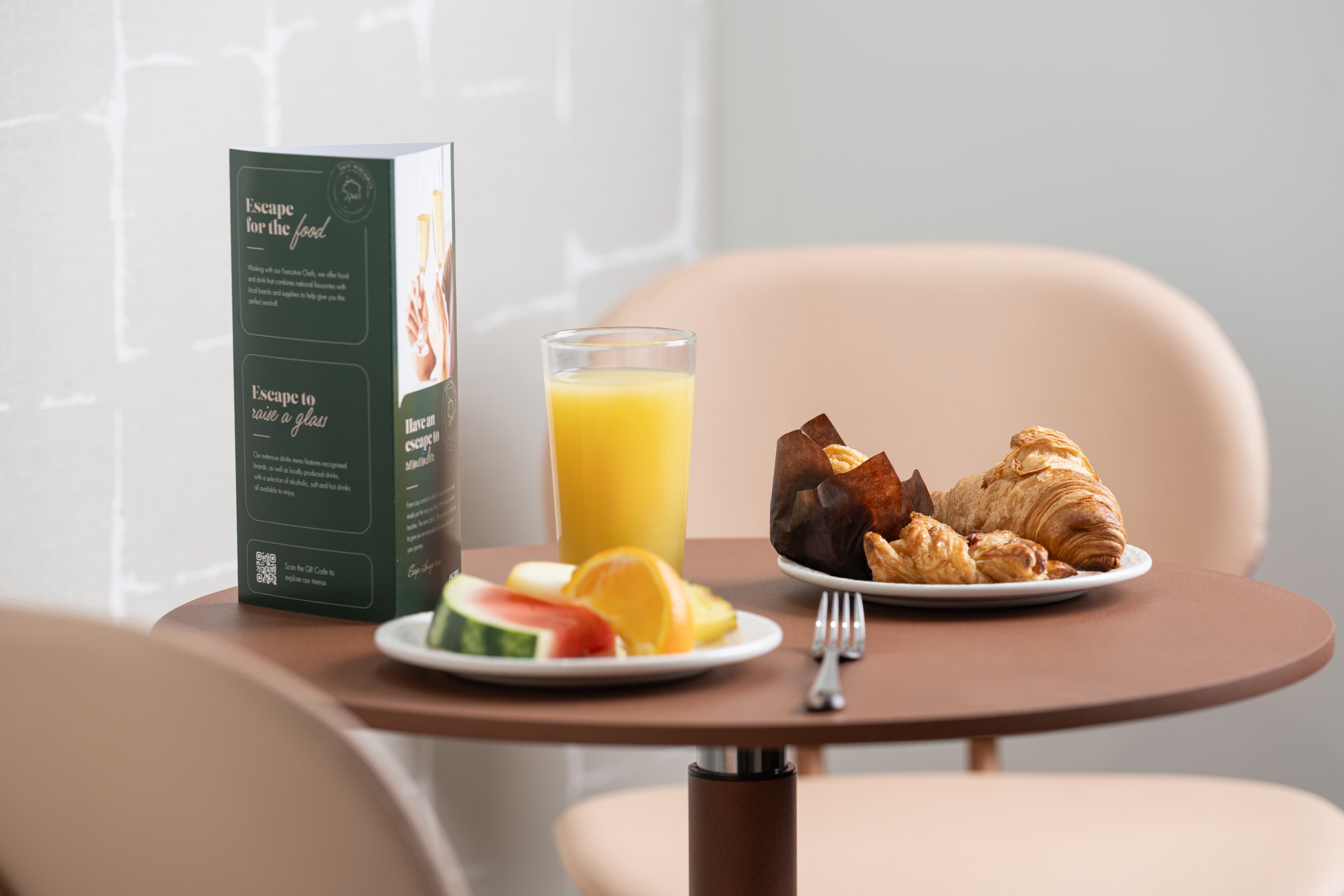 A small table with plates of sliced fruit, croissants, and muffins alongside a glass of orange juice and a menu stand.