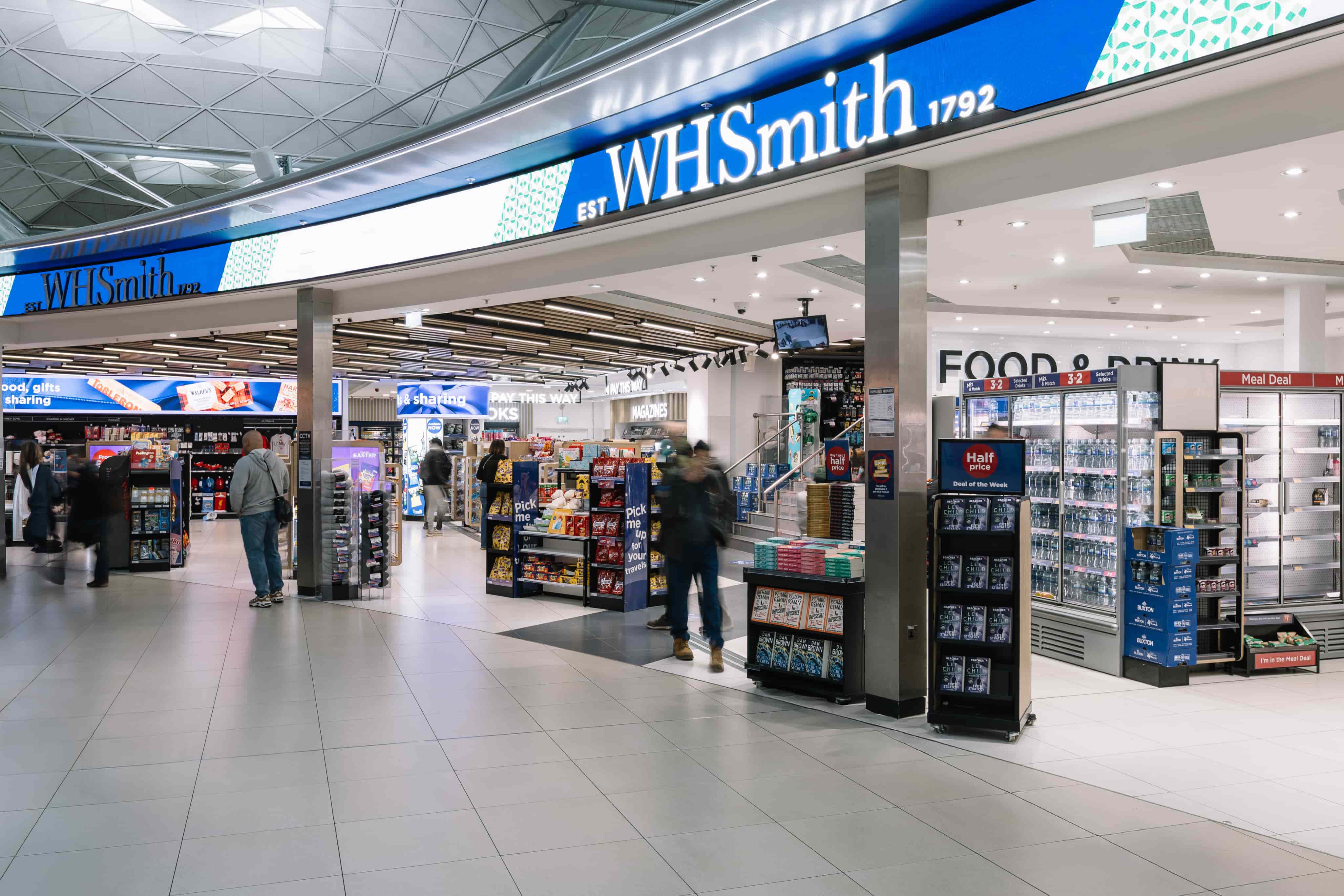 Shoppers browse inside a WHSmith store, displaying books, snacks, and drinks, with bright lighting and a modern design.