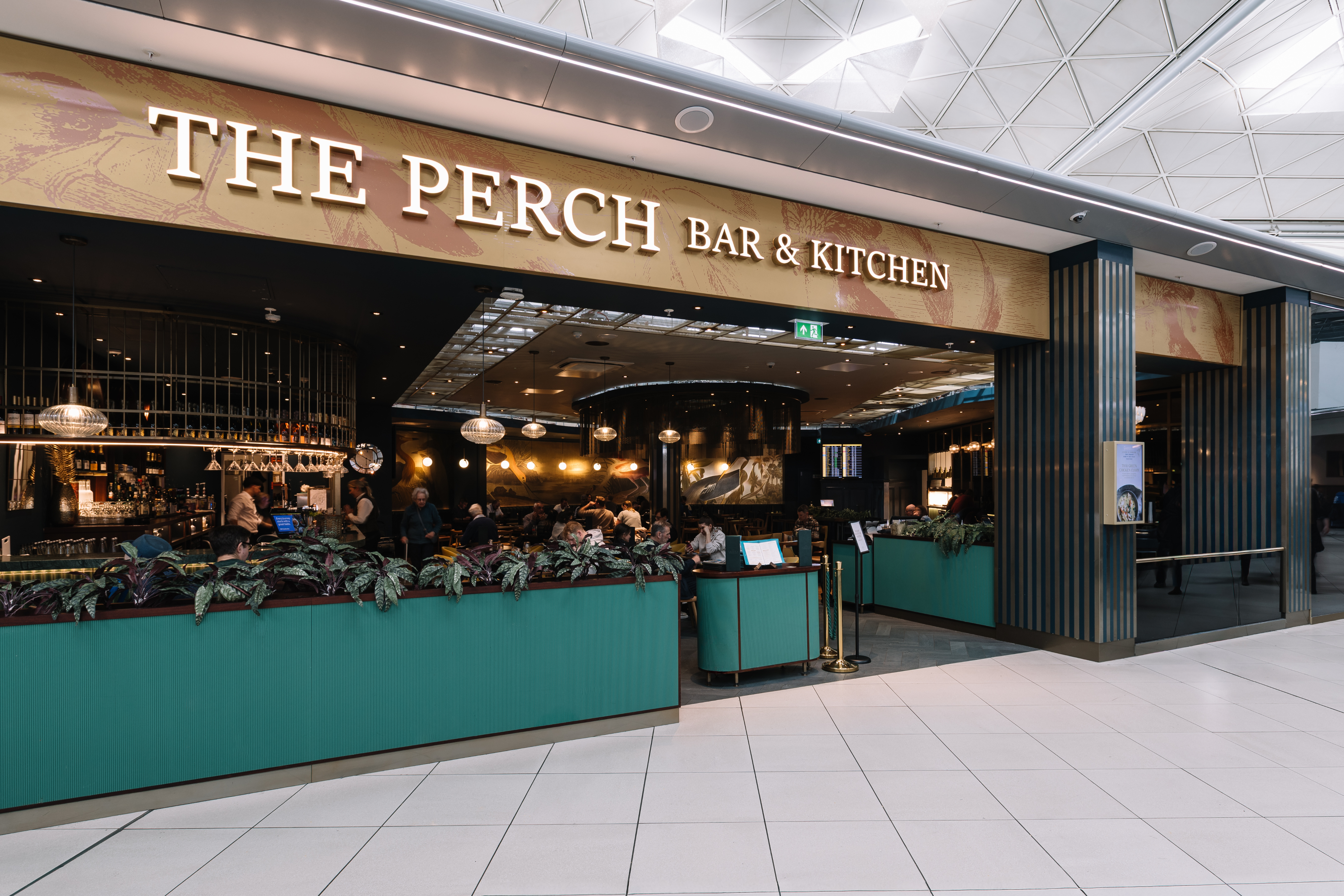 Entrance of The Perch Bar & Kitchen featuring a modern design, hanging lights, and people dining, set in a spacious, tiled airport terminal.