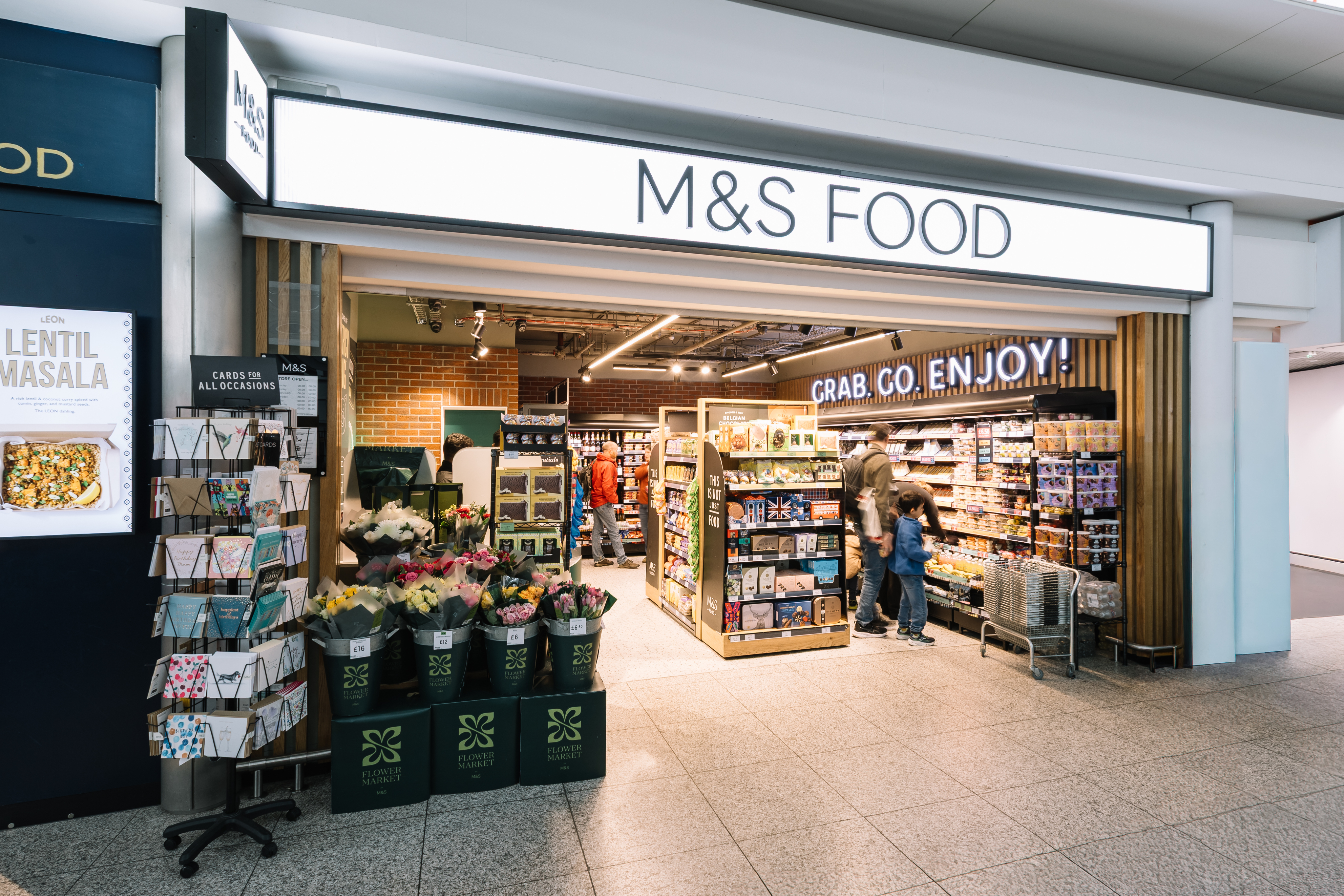 Entrance of M&S Food store with cards, flowers for sale outside; interior shows shelves stocked with groceries.
