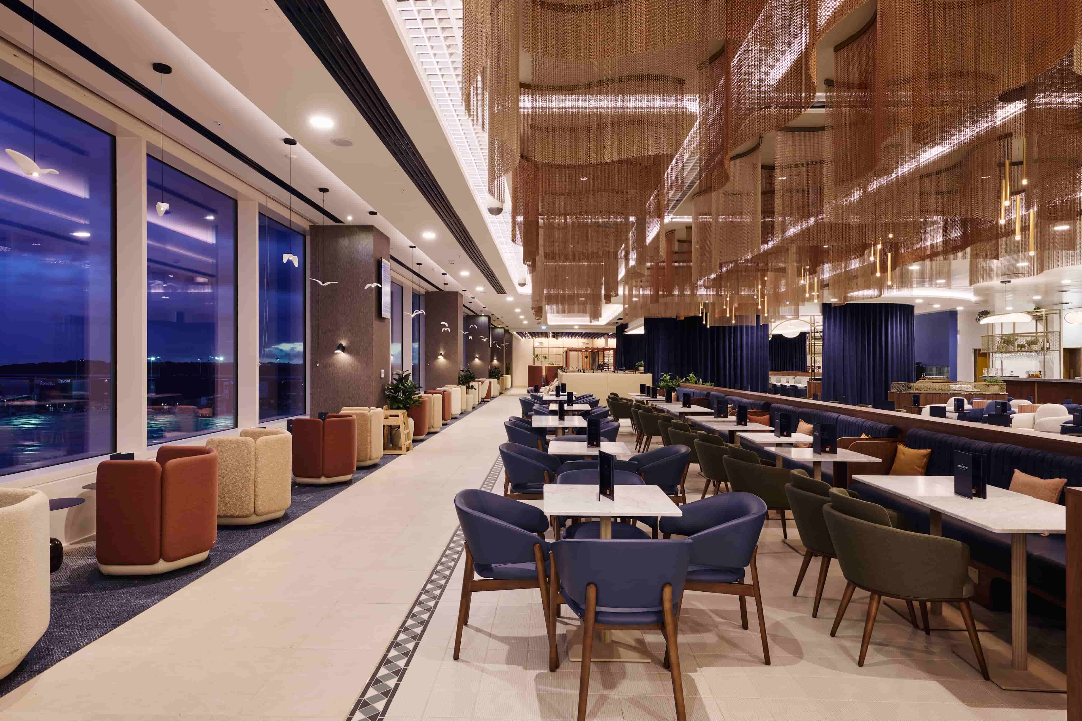 Spacious, modern restaurant interior with purple chairs, white tables, and large windows overlooking the view at dusk. Stylish lighting and decor.