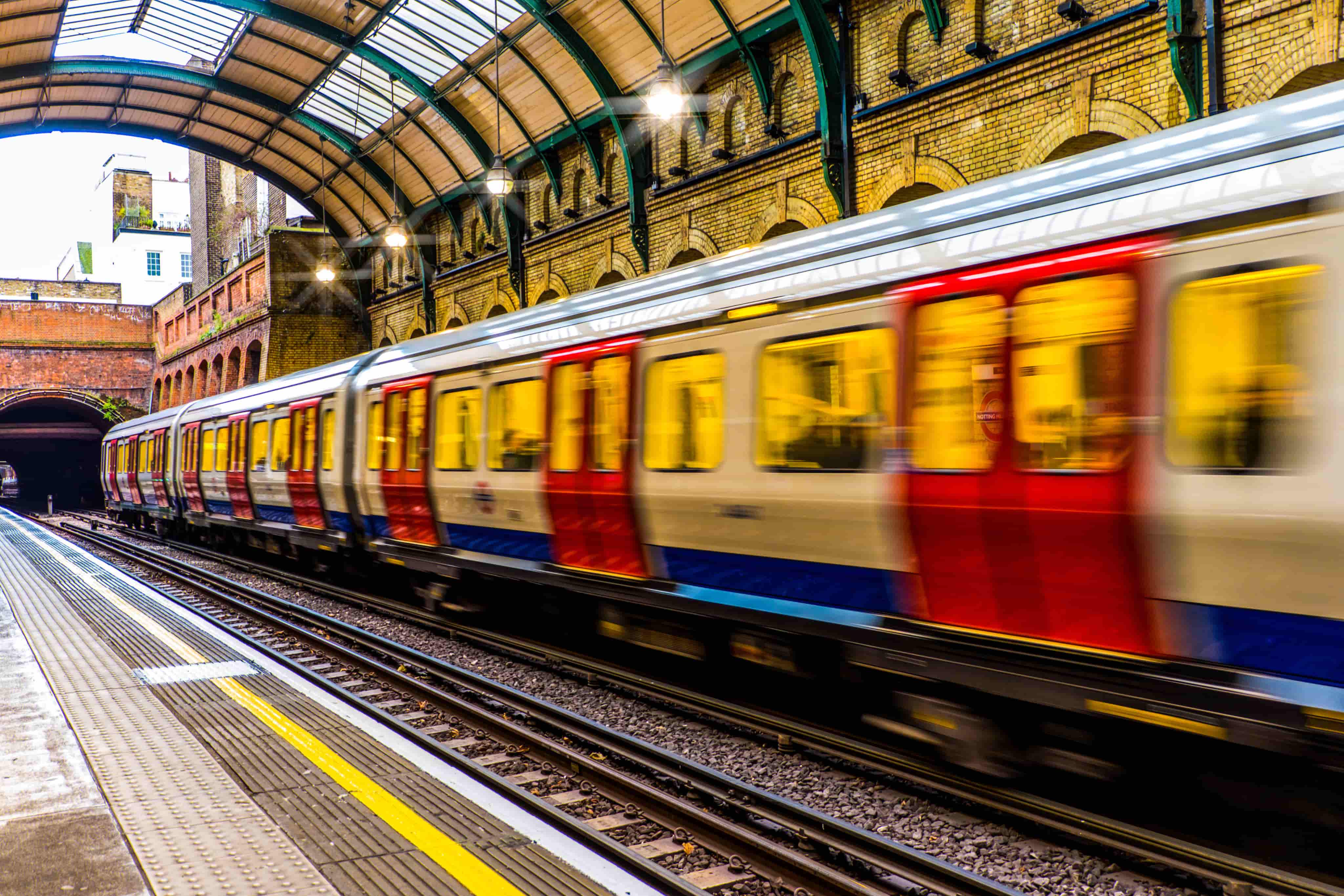 A train speeds through a brightly lit, arched-roof station with brick walls and empty platforms.