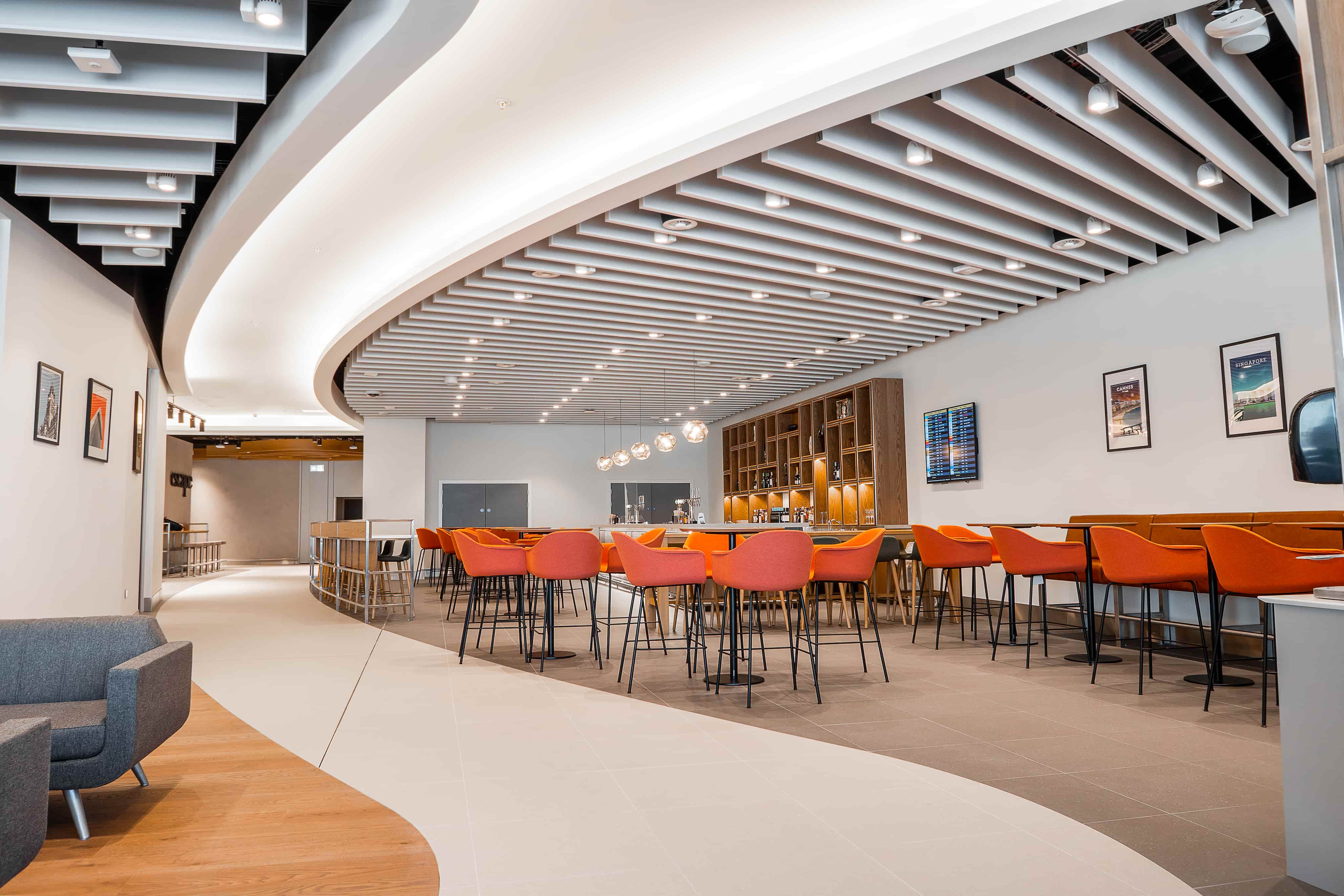 Modern lounge with curved ceiling, orange chairs, wooden tables, and a bar area in a contemporary setting.