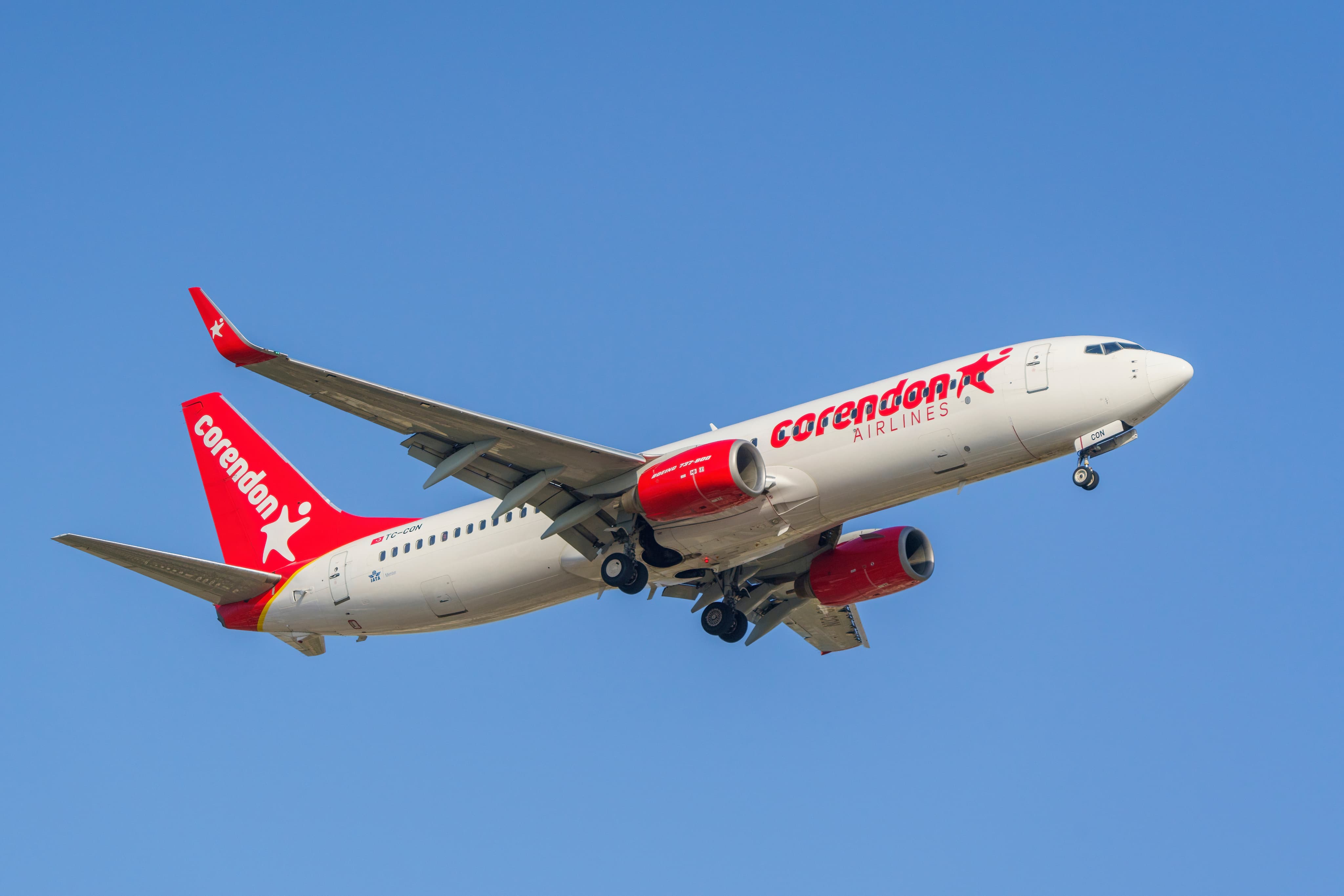 A Corendon Airlines plane with a white body and red accents in flight against a clear blue sky.