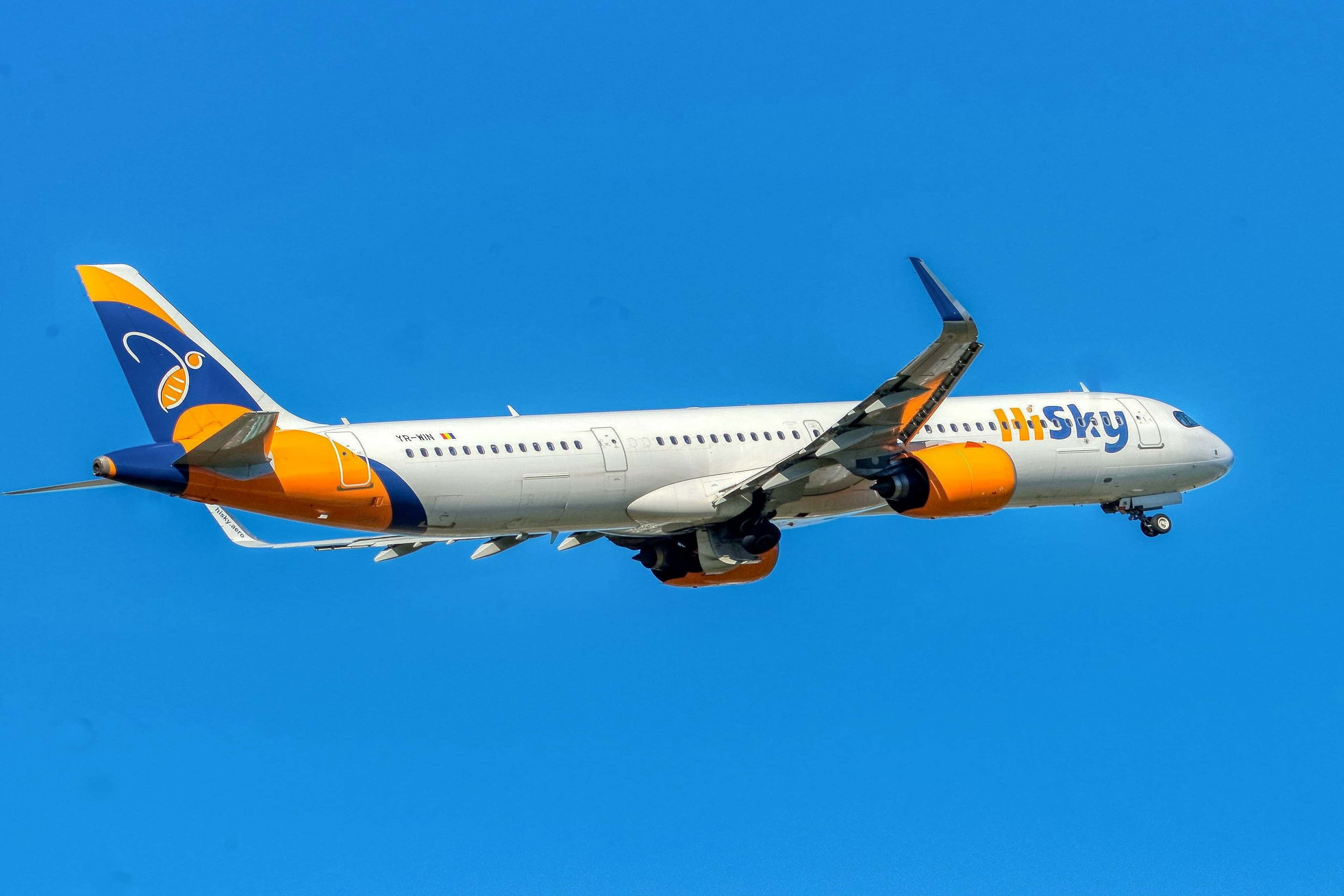 A white airplane with orange and blue accents flying against a clear blue sky.