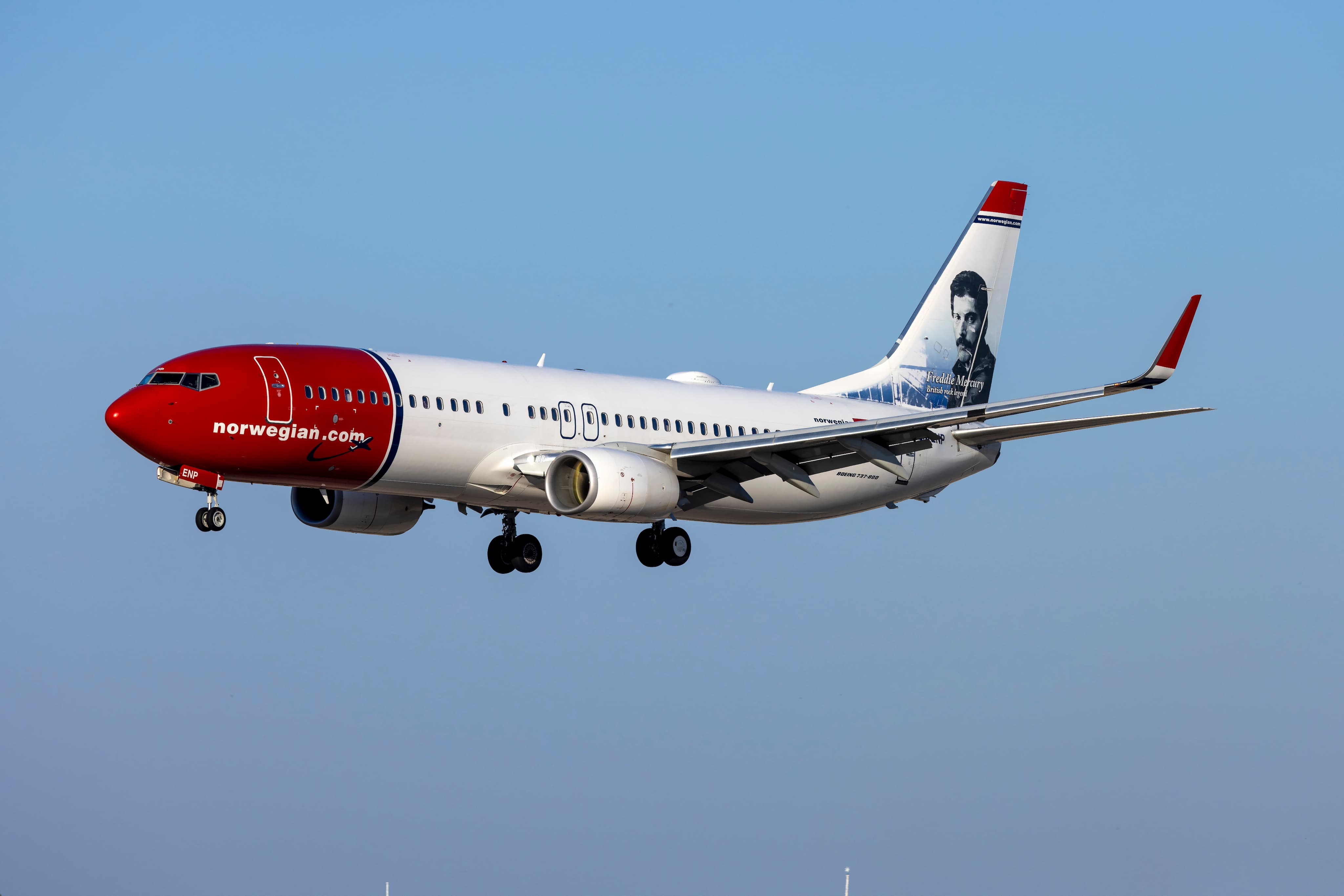 A Norwegian Air plane with a red nose and a portrait on the tail fin, flying against a clear blue sky.