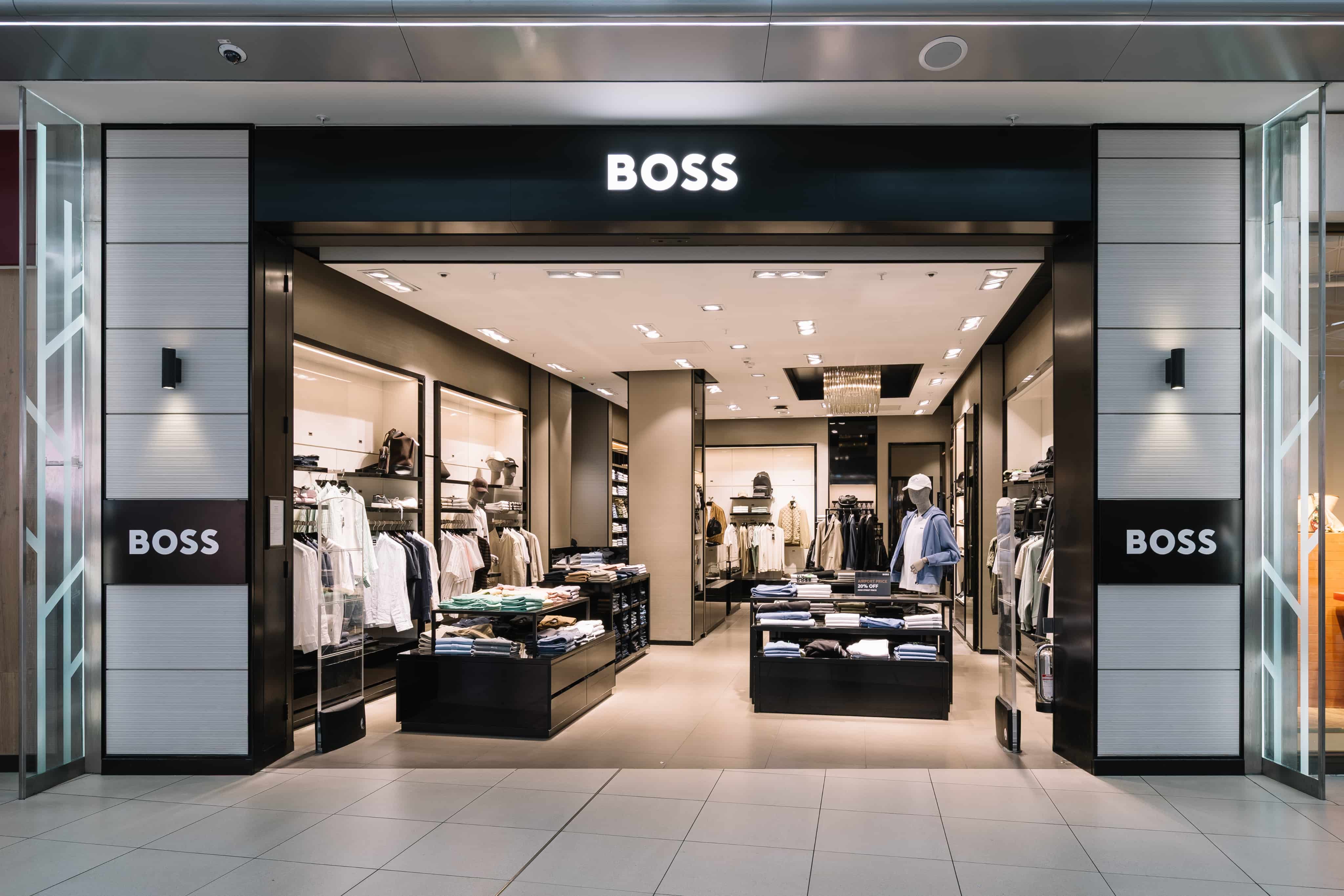 Storefront of a modern clothing store named "BOSS," displaying neatly arranged apparel on tables and mannequins, with stylish interior lighting.